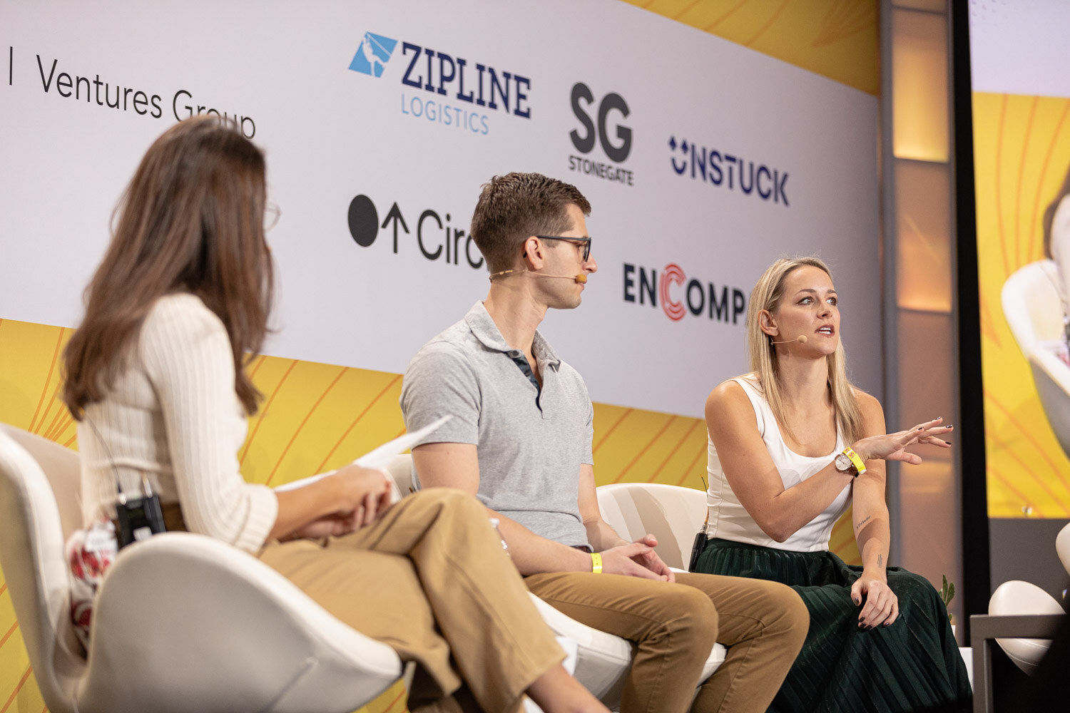 Three people seated on white chairs on stage, the woman on the right gesturing as she speaks.