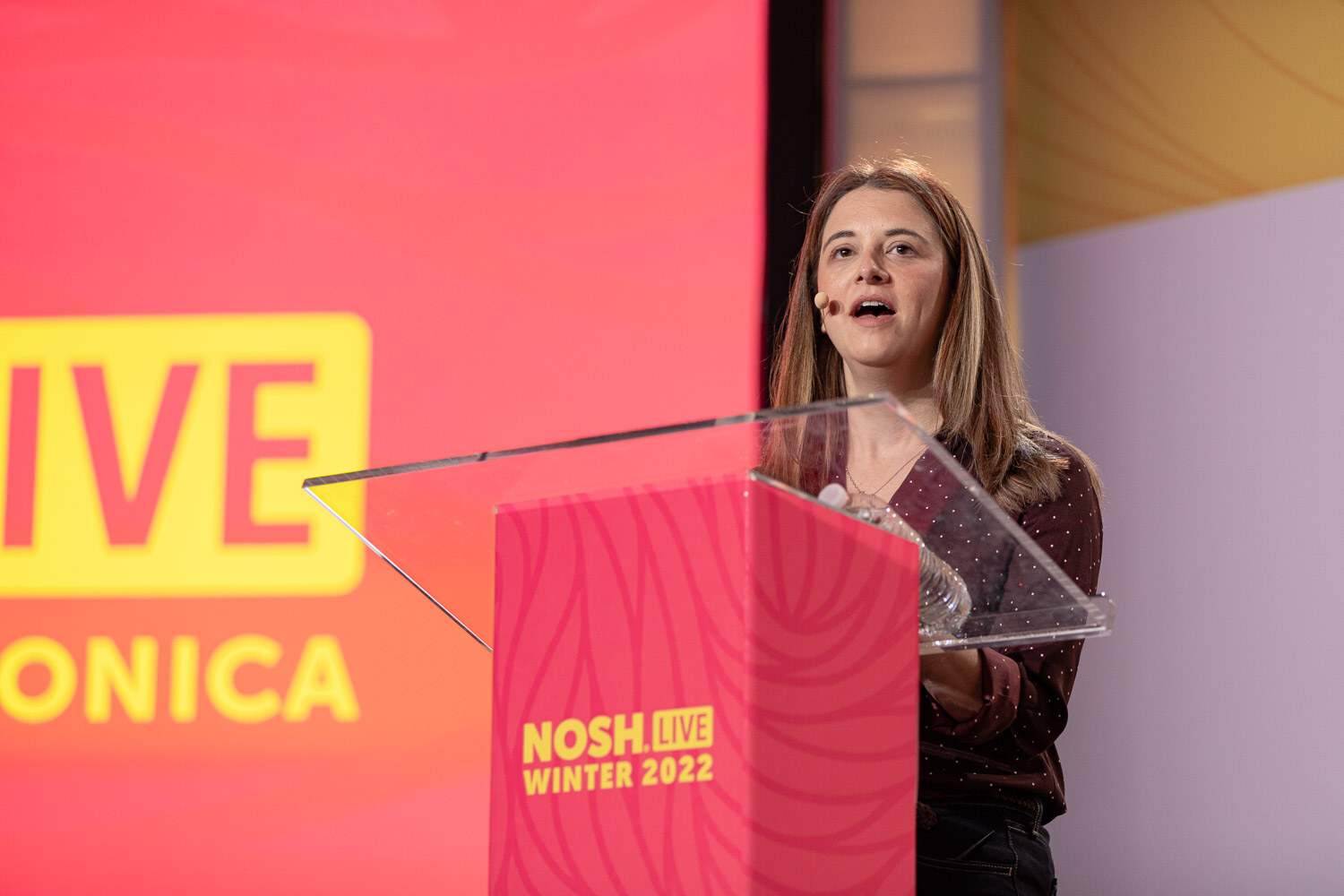 Woman speaking at a pink lectern with a Nosh Live Winter 2022 label.