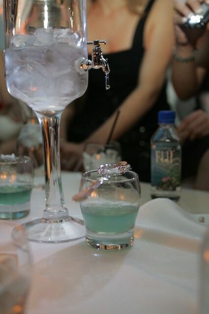 A tall glass dispenser filled with ice and fitted with a tap stands beside a glass of pale green drink. A FIJI water bottle sits behind, with a woman in a black dress and a hand holding a camera.