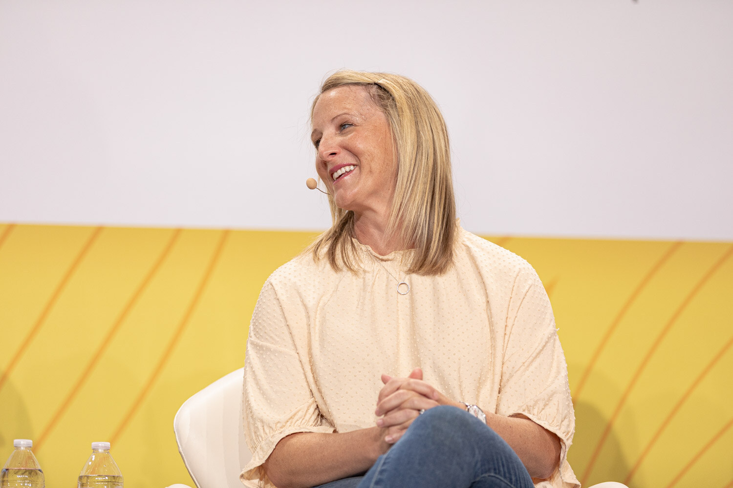 Blond woman in a cream blouse with a headset microphone smiling on a stage with a yellow backdrop.