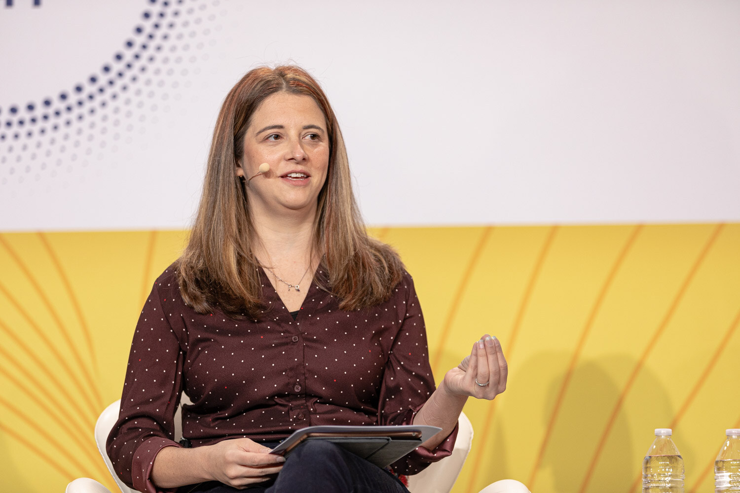 Woman in a burgundy polka-dot blouse holding a tablet and gesturing with one hand.