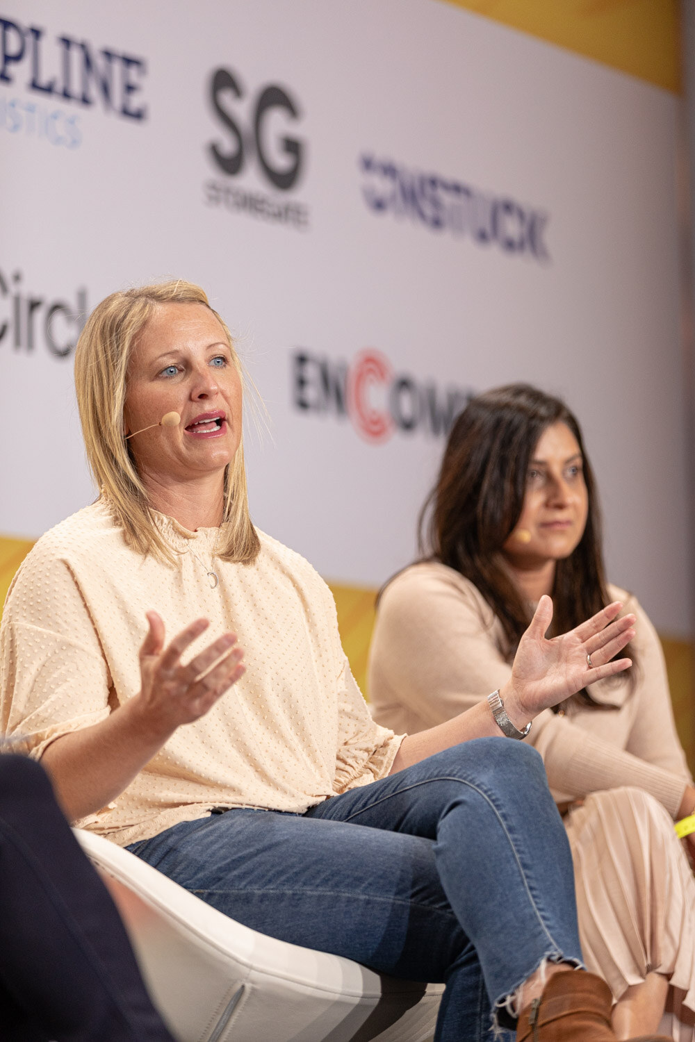 Woman in a cream blouse gesturing next to a woman in a beige top, seated in front of sponsor logos.