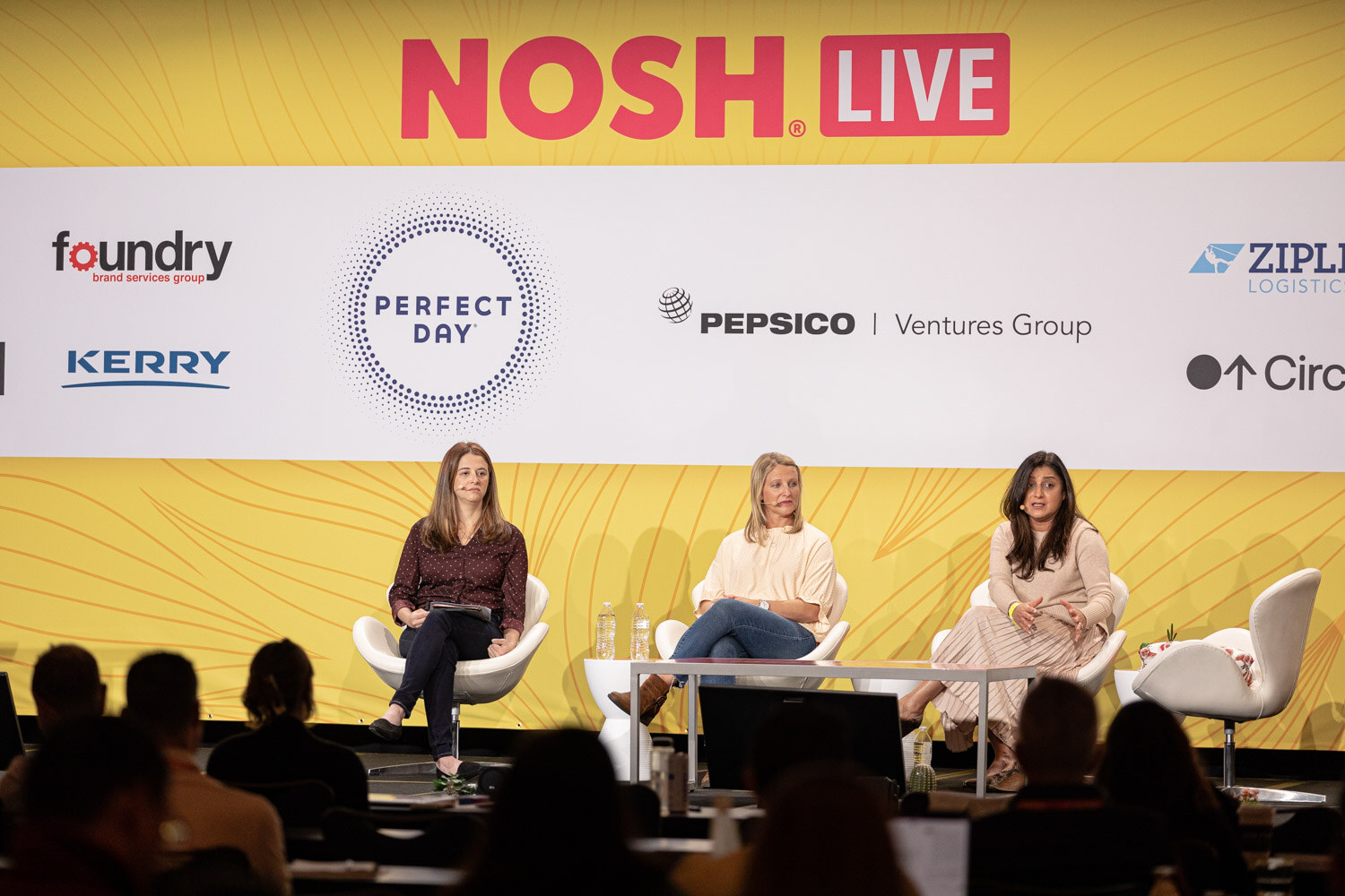 Wide shot of three women seated on a stage with a Nosh Live banner and audience silhouettes in front.
