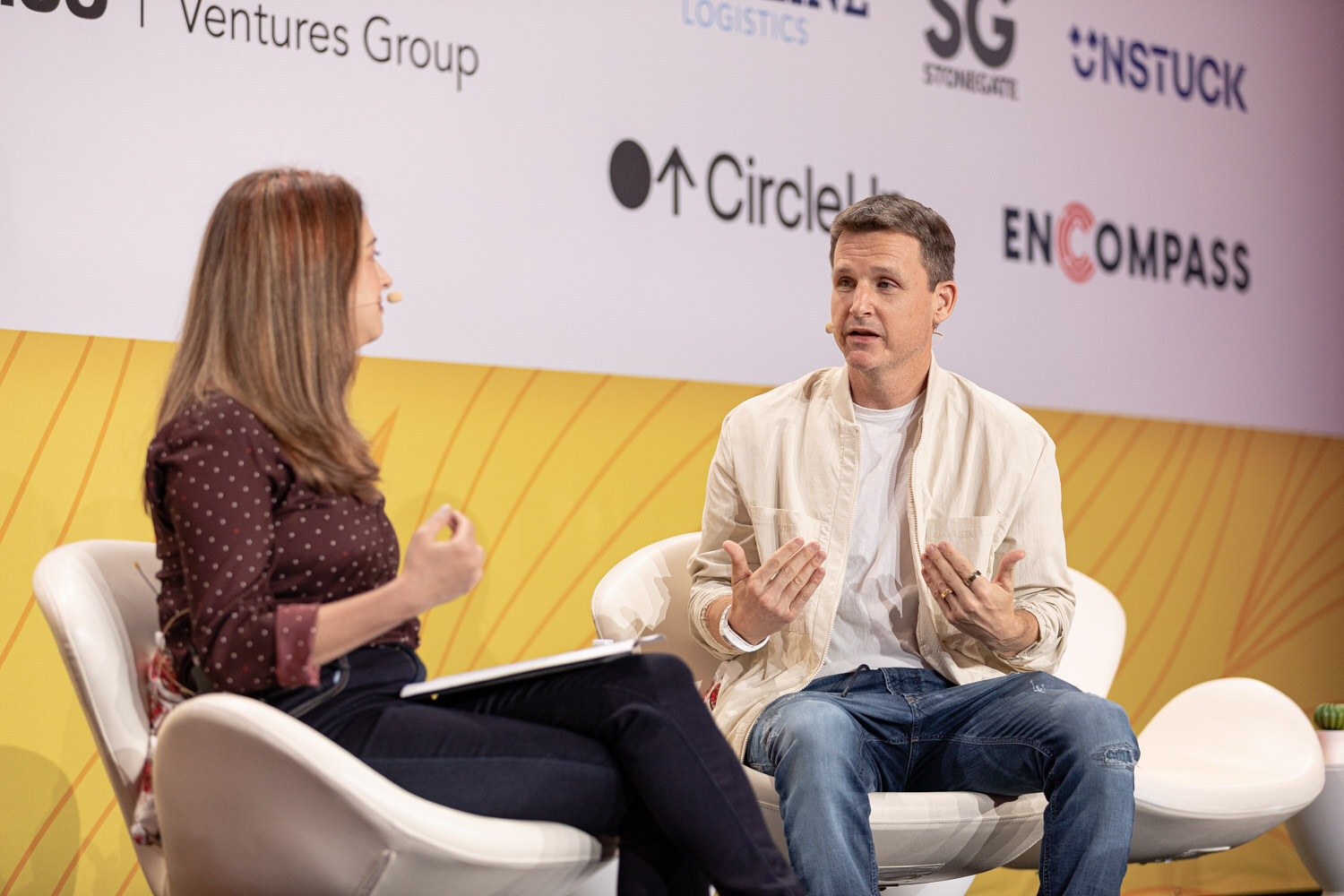 Woman in a polka-dot top and man in a cream jacket seated and talking in front of Pepsico Ventures Group and other sponsor logos.