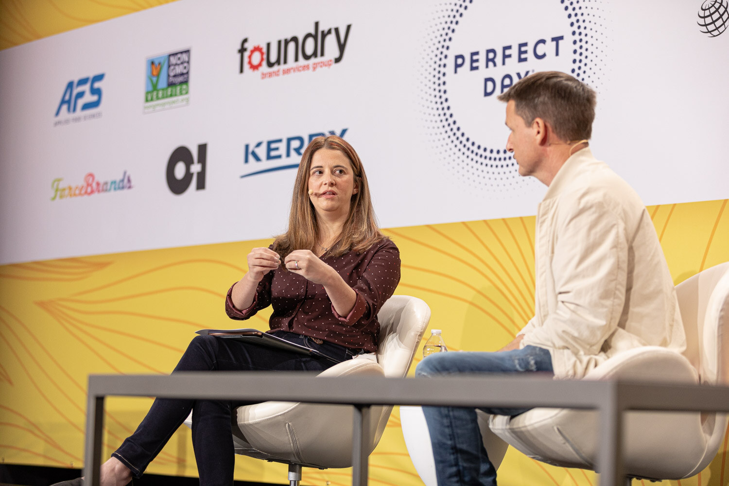 Woman with a notepad seated on stage facing a man in a cream jacket, sponsor logos behind them.