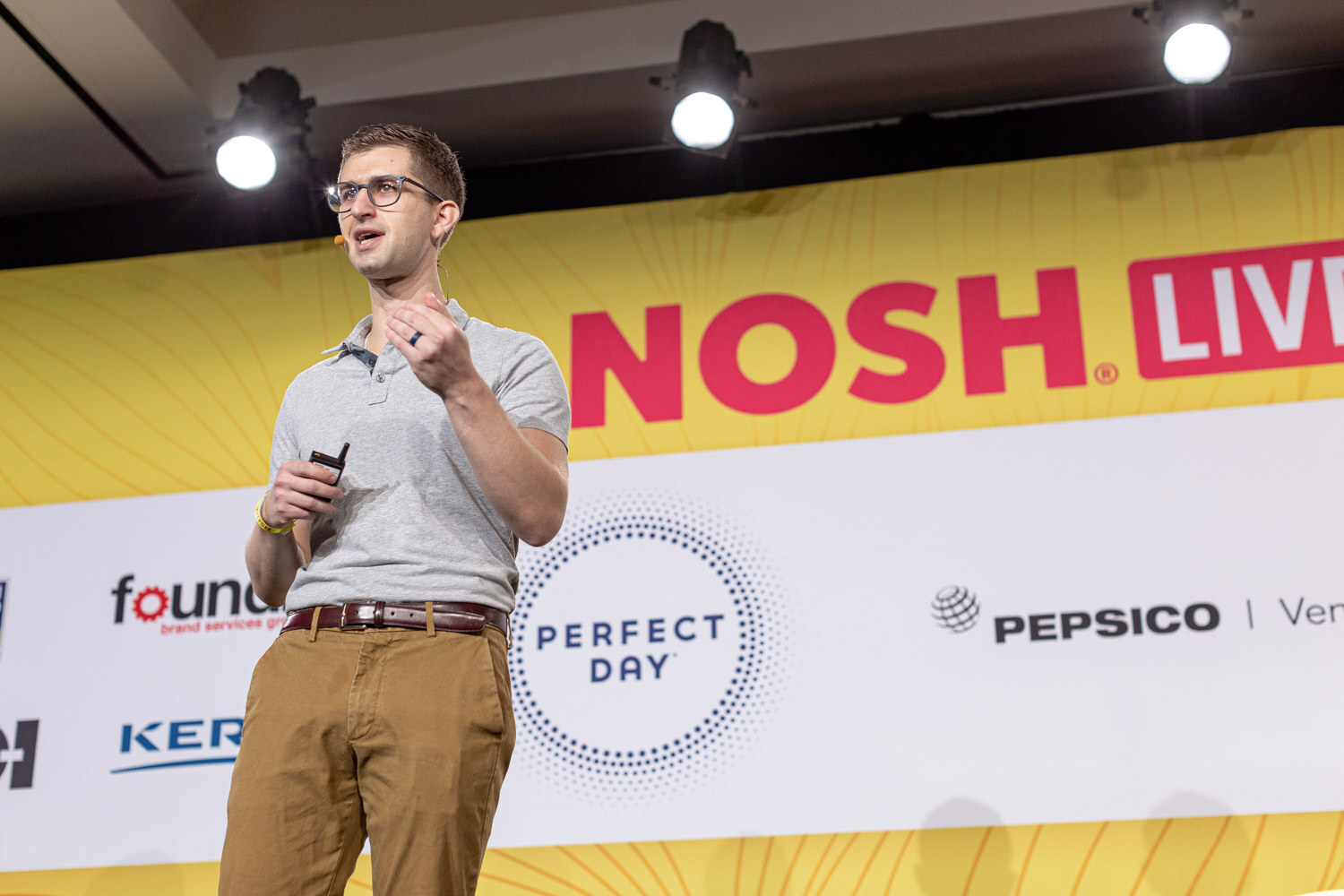 Man in a gray polo and khakis speaking on stage in front of a Nosh Live banner.