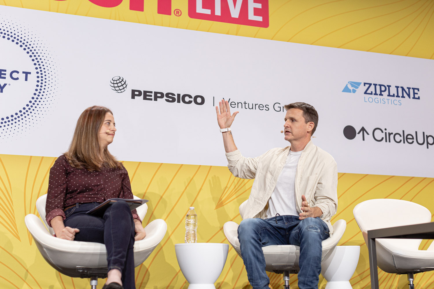 Man in a cream jacket with an arm in the air next to a woman holding a tablet, on a stage with sponsor logos.