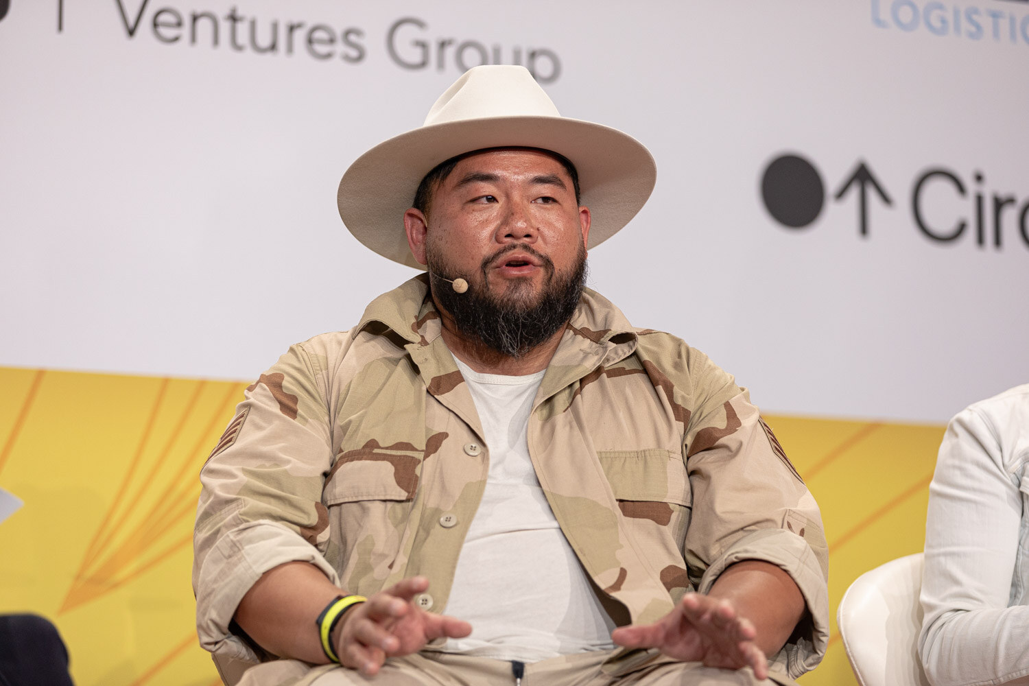 Bearded man in a cream hat and tan camo jacket speaking, hands out in front of him.