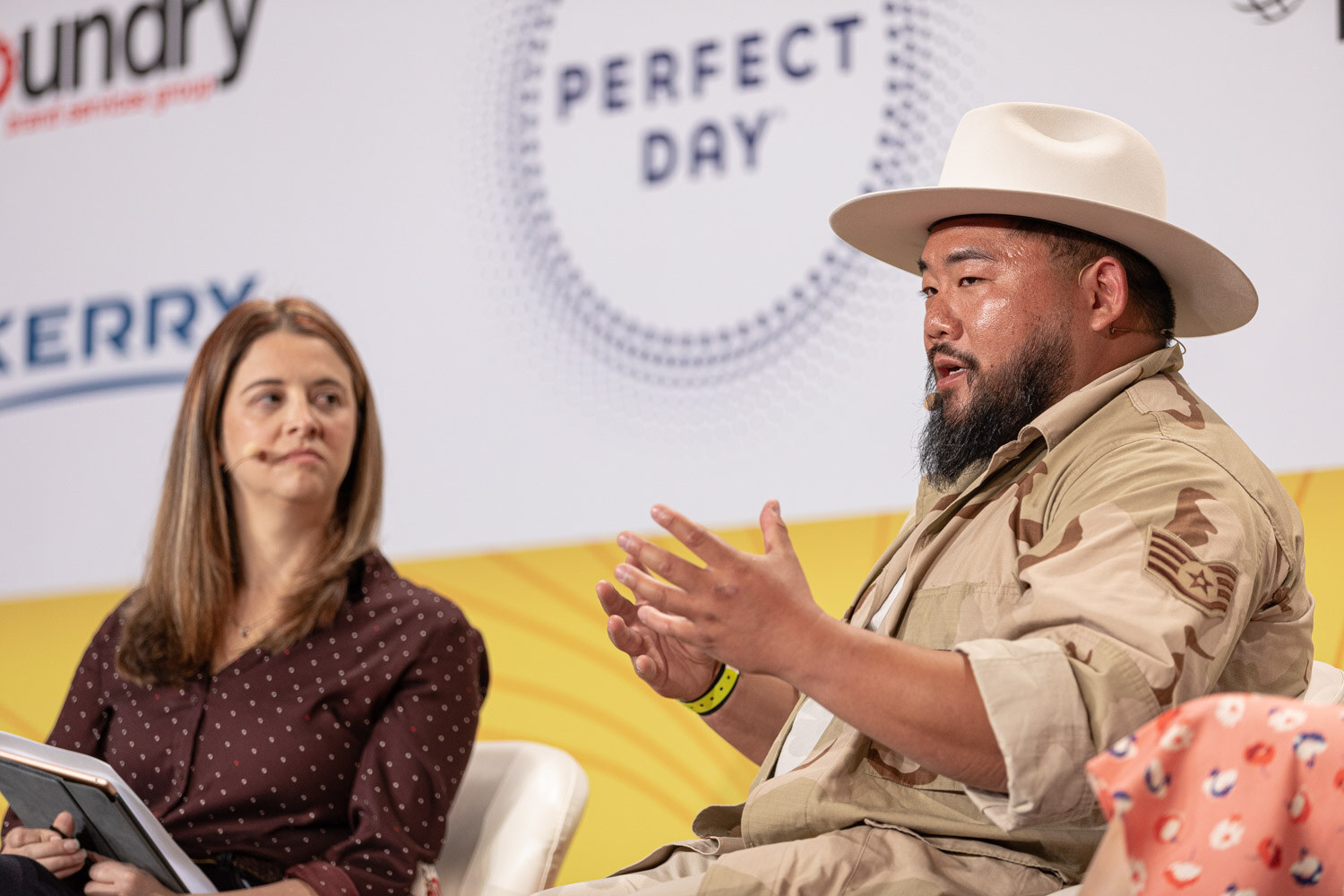 Man in a cream hat gesturing with both hands while a woman in a polka-dot top listens.