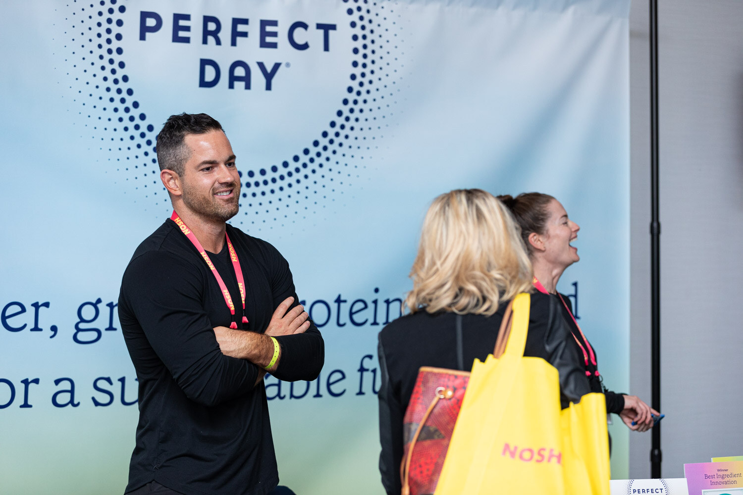 Man in a black shirt with arms crossed beside a Perfect Day banner, two women in conversation next to him.