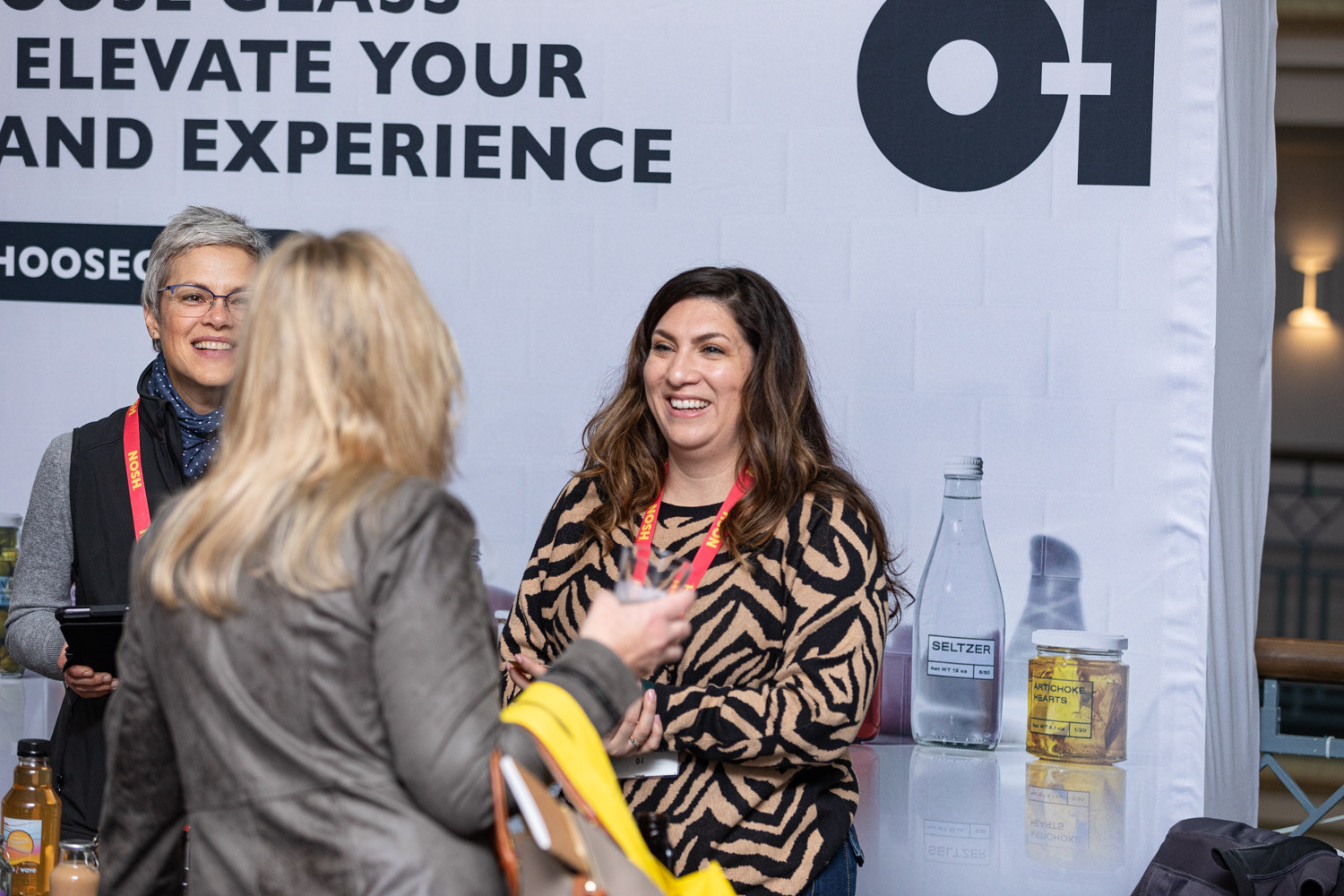 Woman in a tiger-print top smiling at a display of glass bottles and jars, with an O-I banner behind her.
