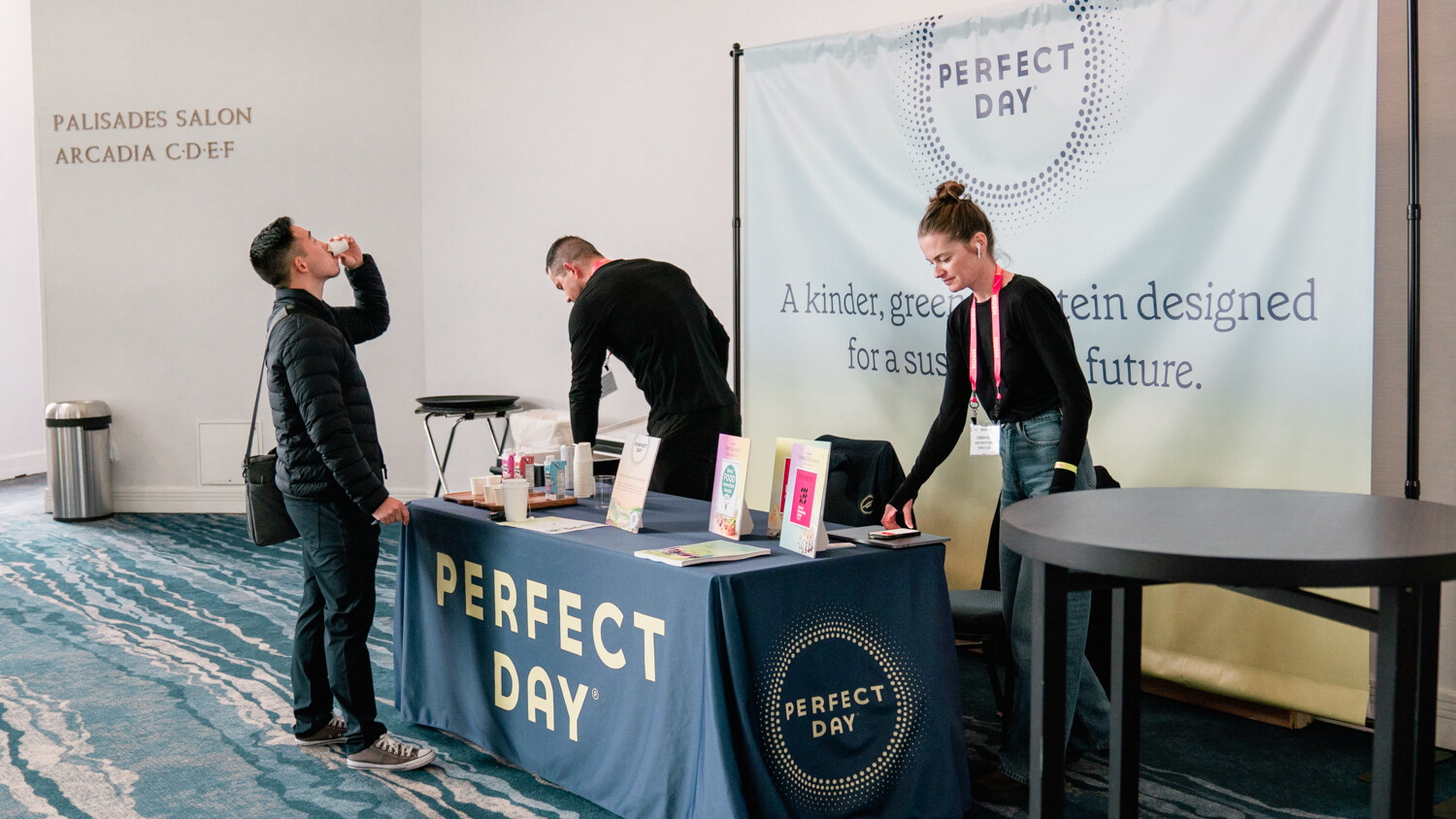 A man drinking a sample next to a Perfect Day table with a woman and a man behind the table.