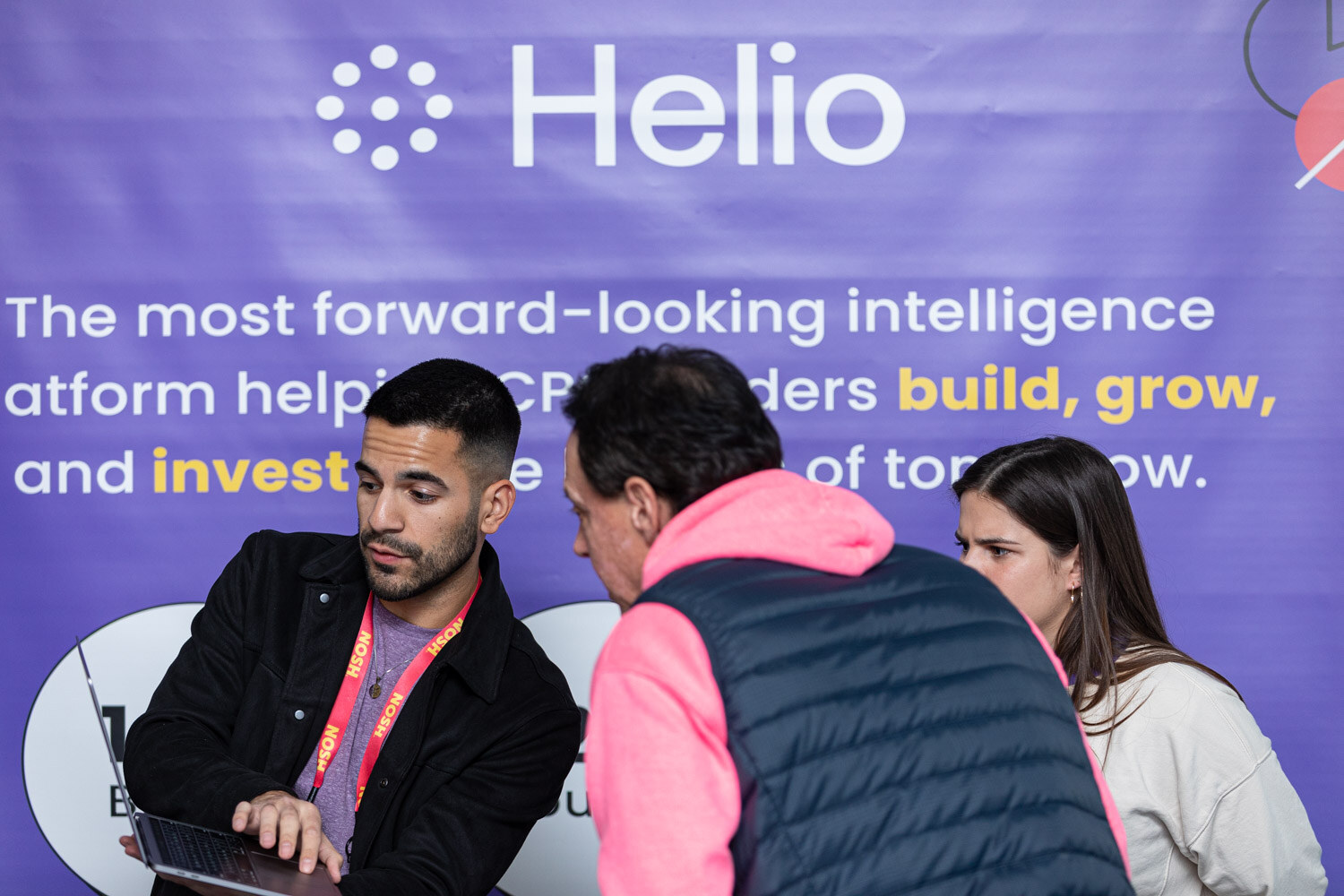 Two men leaning over a laptop in front of a purple Helio banner, with a woman behind them.