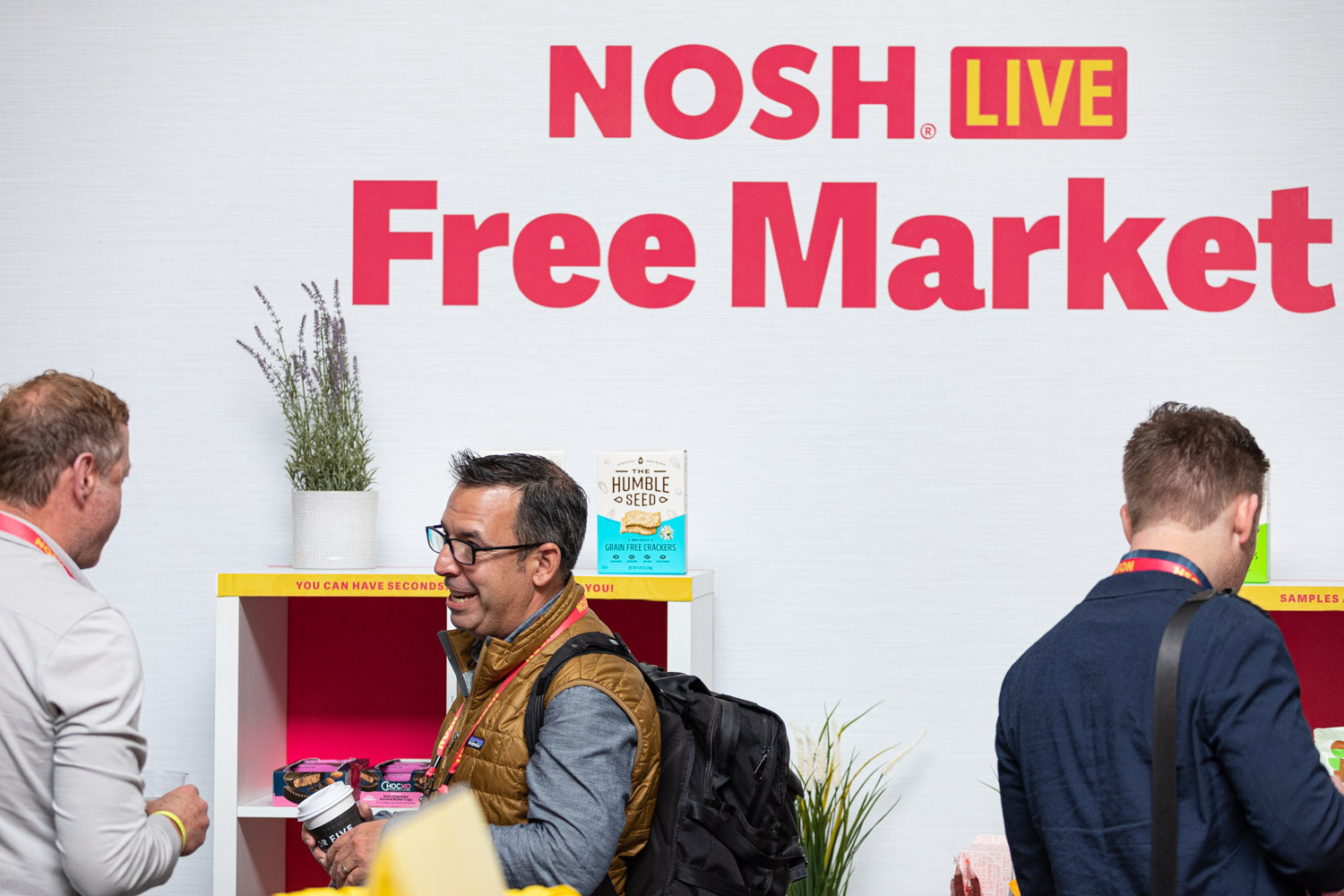 Men standing in front of a Nosh Live Free Market sign and a shelf of snack samples.