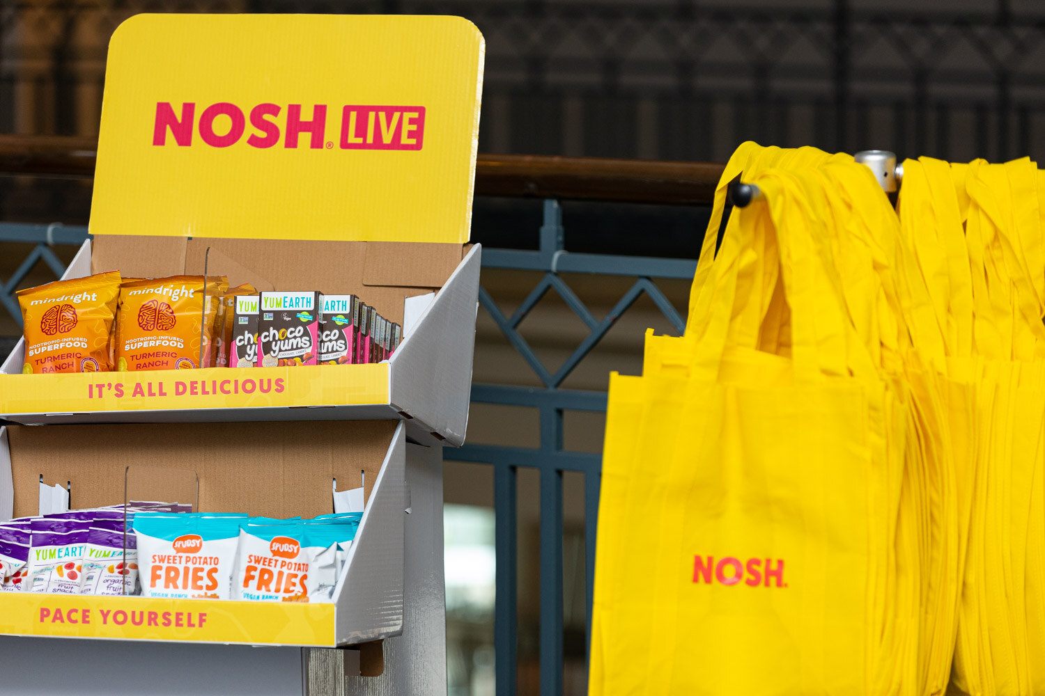 Yellow cardboard display shelves with snack packages next to a rack of yellow Nosh tote bags.