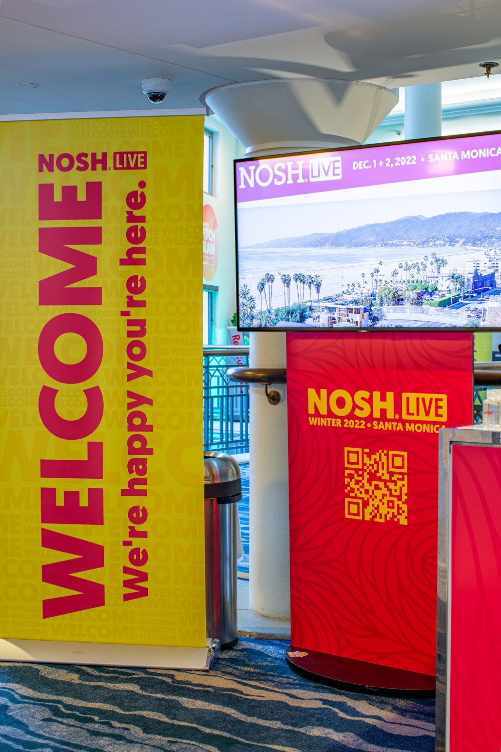Yellow Welcome banner beside a pink pedestal and a screen showing a Santa Monica coastline and the dates of the event.