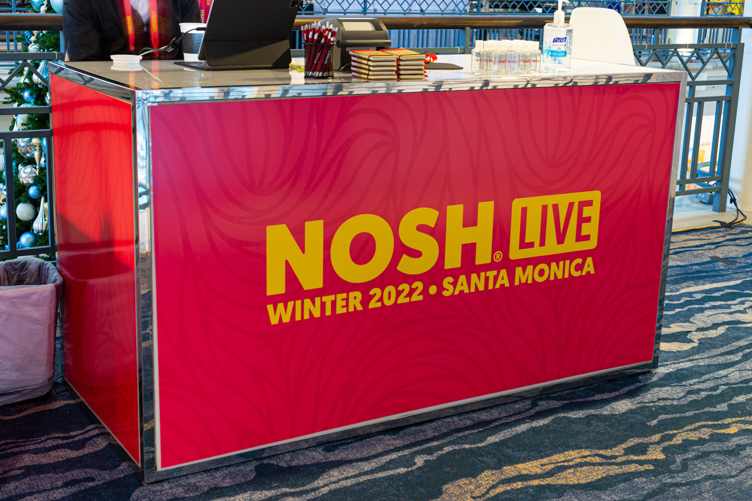 Pink counter with yellow text reading Nosh Live Winter 2022 Santa Monica, with a laptop and supplies on top.