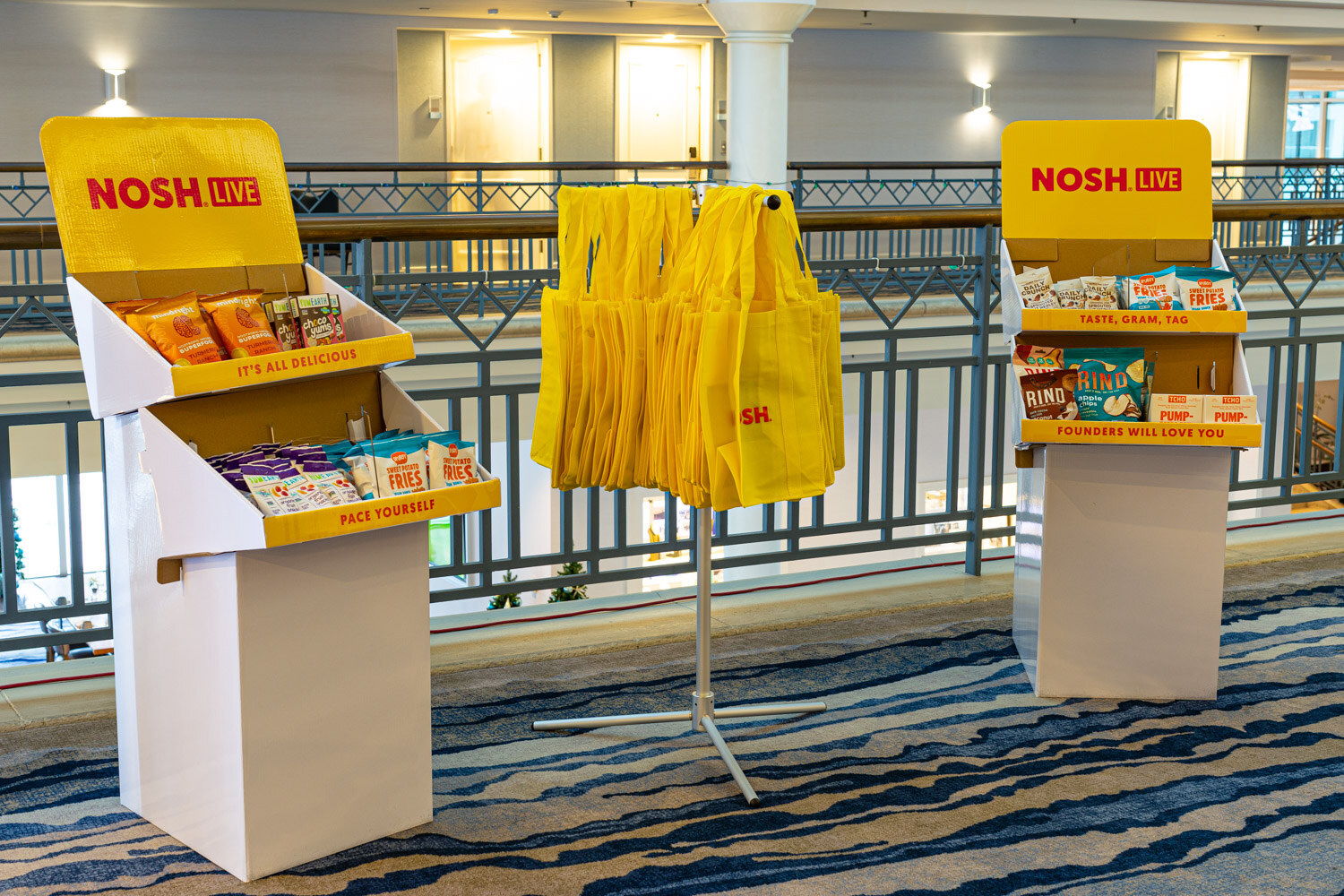 Two yellow Nosh Live display stands with snacks and a rack of yellow tote bags beside a balcony railing.