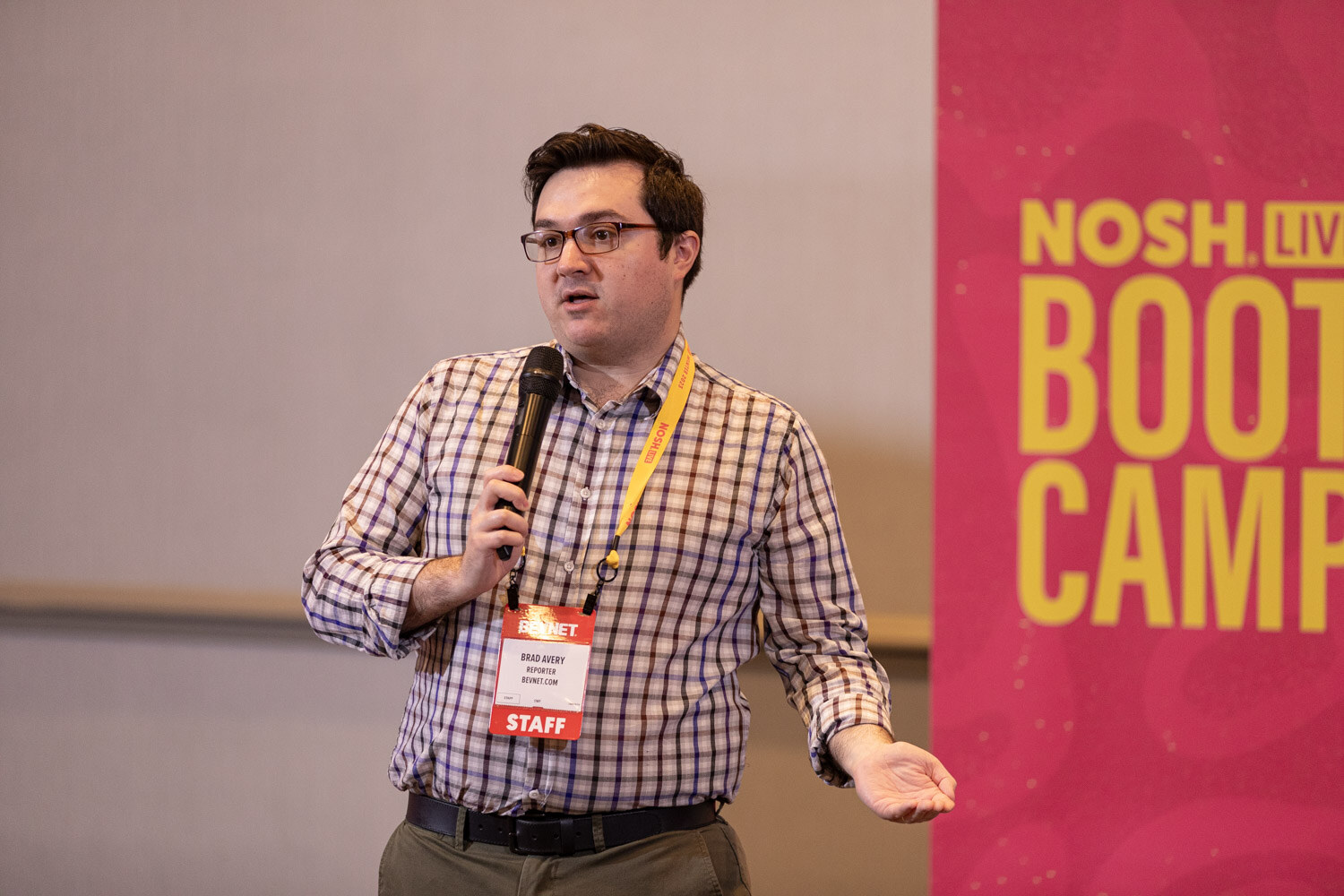 A man holding a microphone next to a yellow and pink NOSH LIVE BOOT CAMP banner.