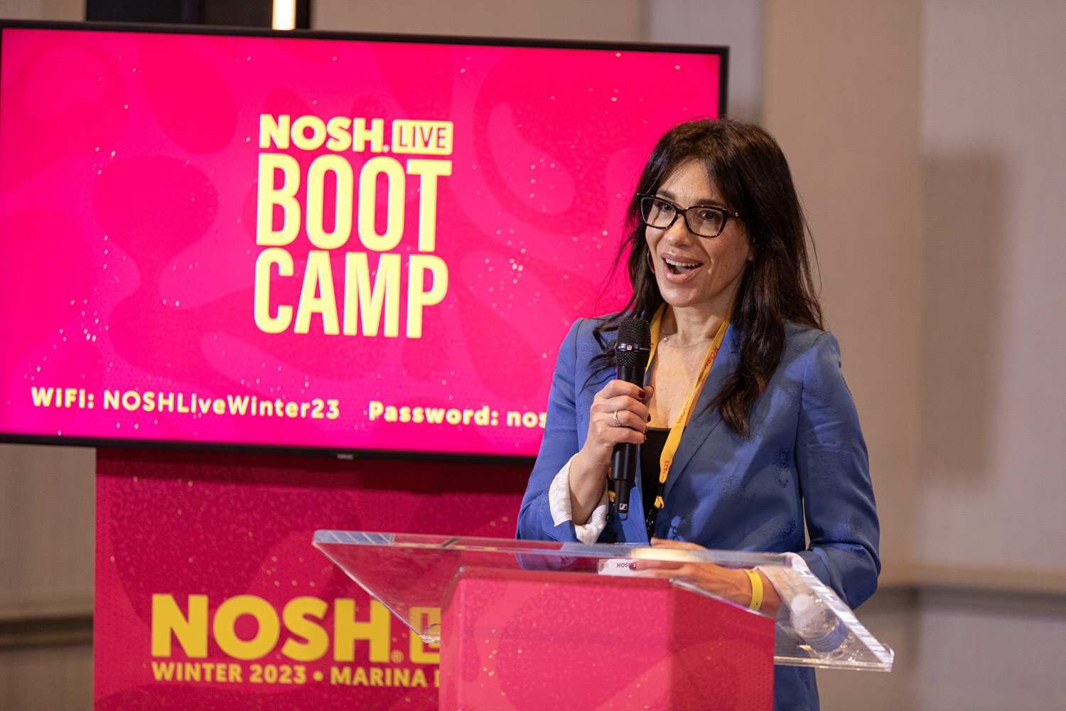 A woman with glasses speaking at a podium beside a screen reading NOSH LIVE BOOT CAMP.