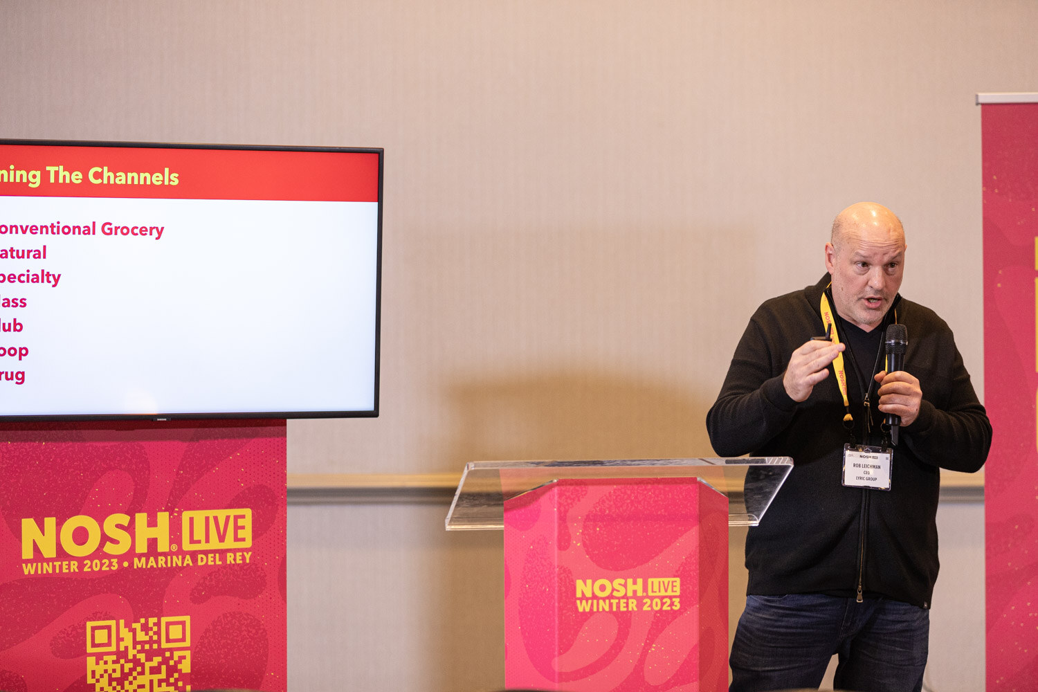 A man at a podium beside a screen with a slide listing conventional grocery, natural, specialty and other channels.
