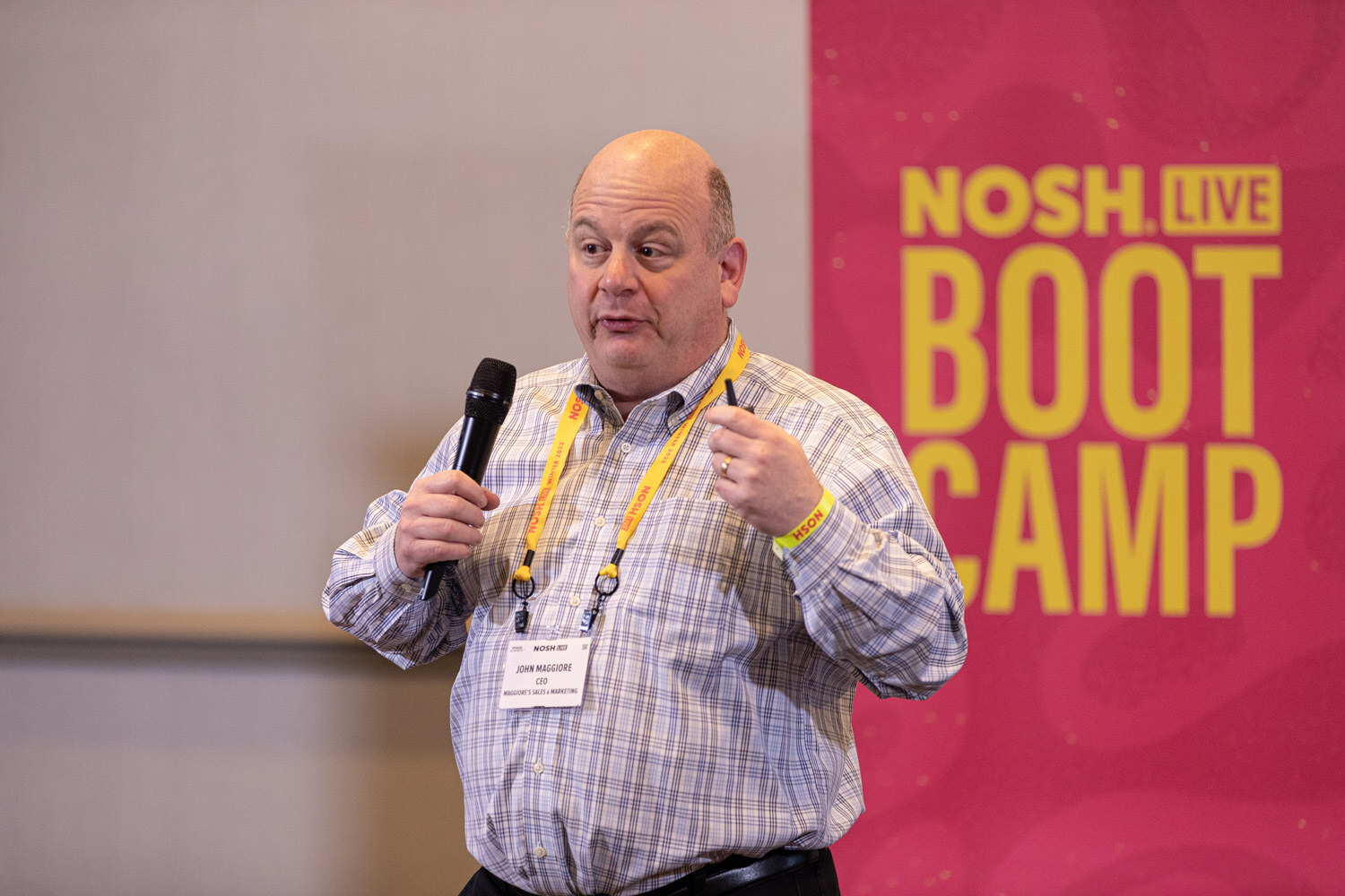 A man in a checked shirt holding a microphone in front of a NOSH LIVE BOOT CAMP banner.