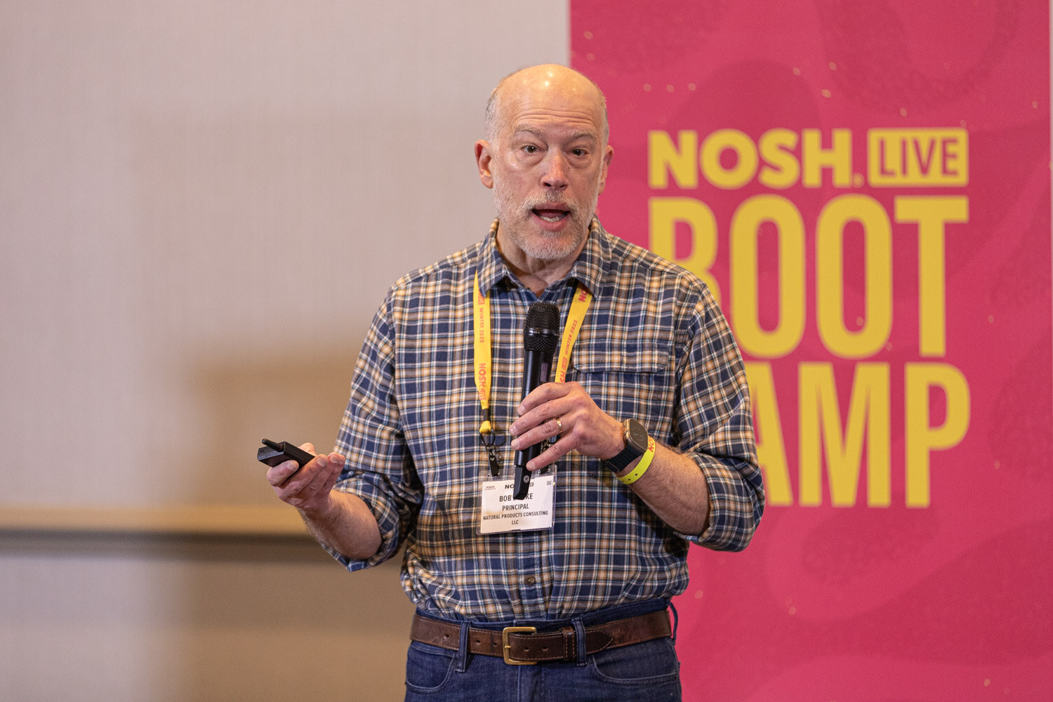 A man in a plaid shirt with a lanyard speaking in front of a Boot Camp banner.