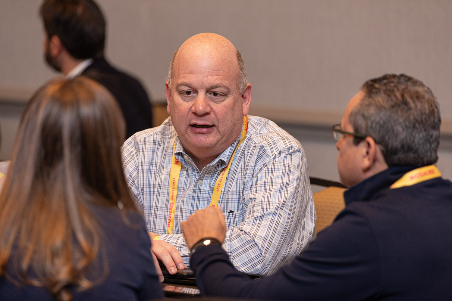 A man with a lanyard talking with others at a small round table.