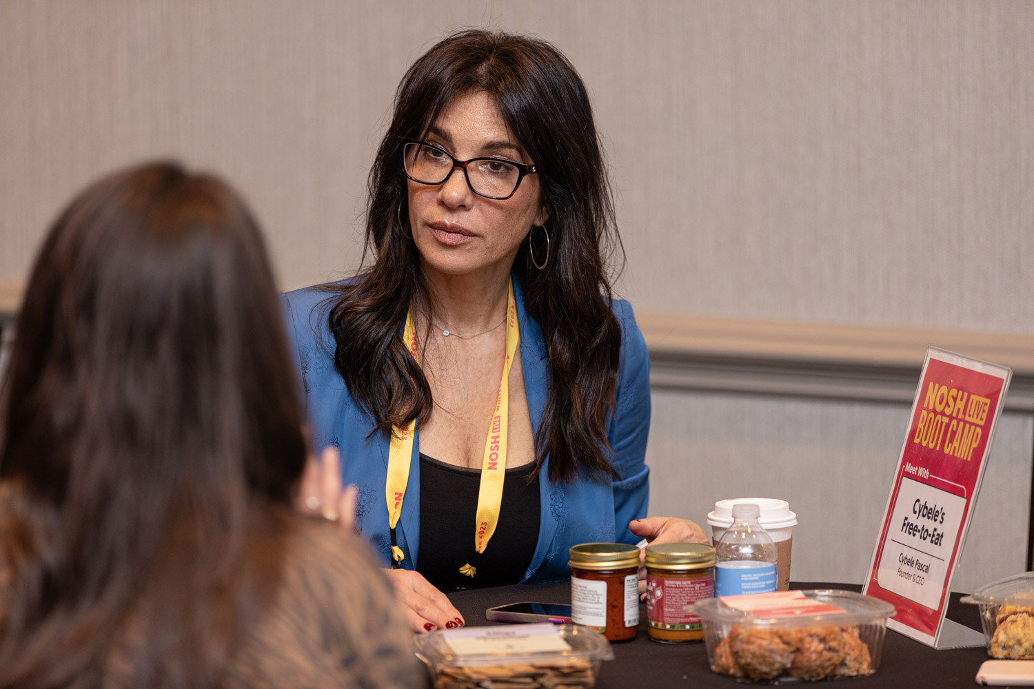 A woman in a blue blazer talking at a table with a Cybele's Free-to-Eat sign and jars of samples.