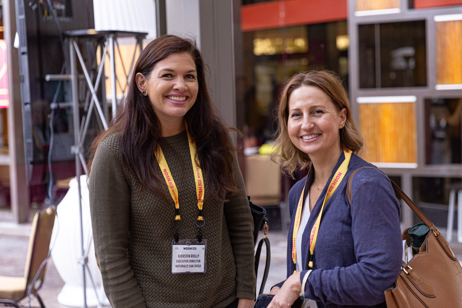 Two smiling women wearing Nosh Live lanyards in a hallway.