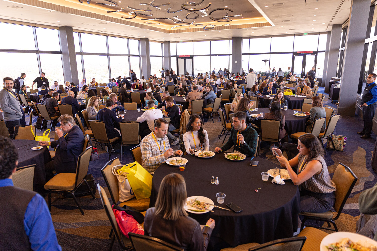 Many attendees seated at round tables with plates in a ballroom with a decorative ceiling.