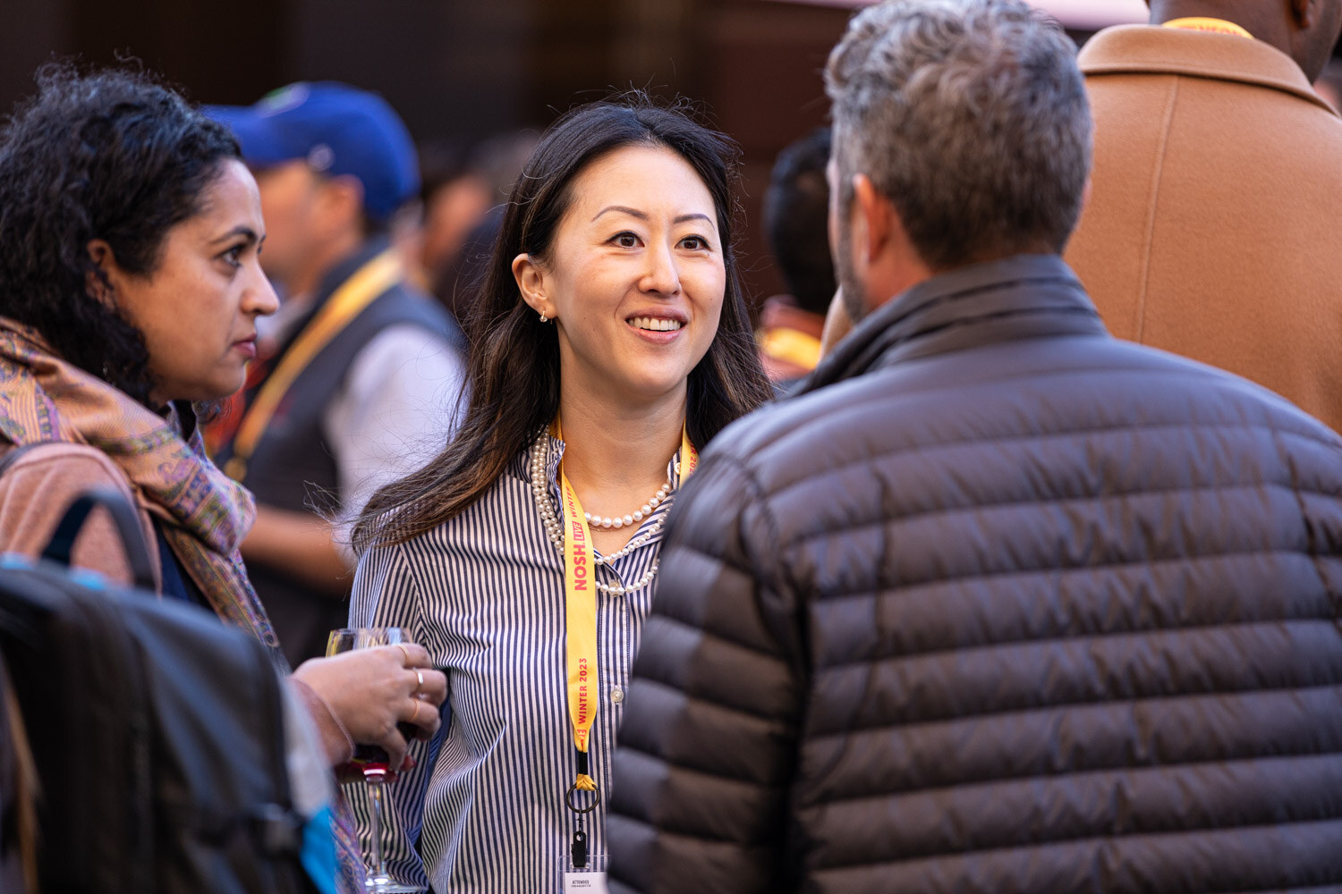 A smiling woman with a lanyard and a glass of wine talking to a man in a puffer jacket.