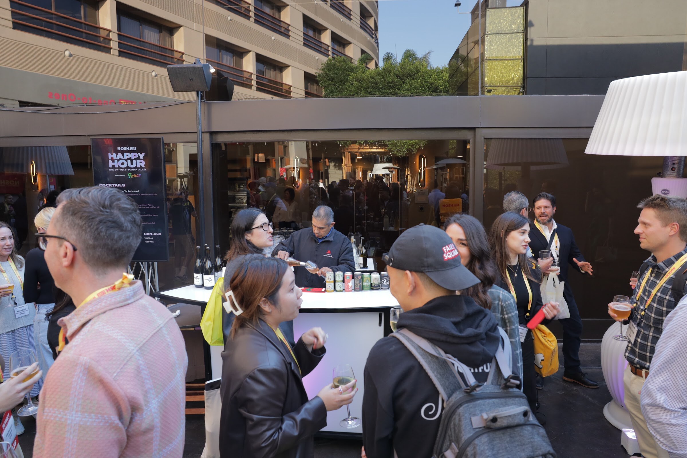 Attendees gathered around a tent bar with a Happy Hour sign in an outdoor courtyard.