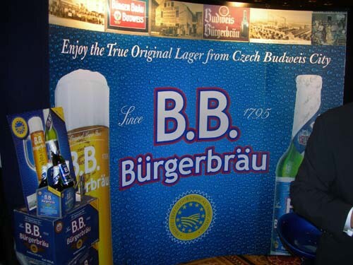 A blue B.B. Bürgerbräu backdrop reads Enjoy the True Original Lager from Czech Budweis City, with stacked product boxes at left and a person in a dark suit at right.