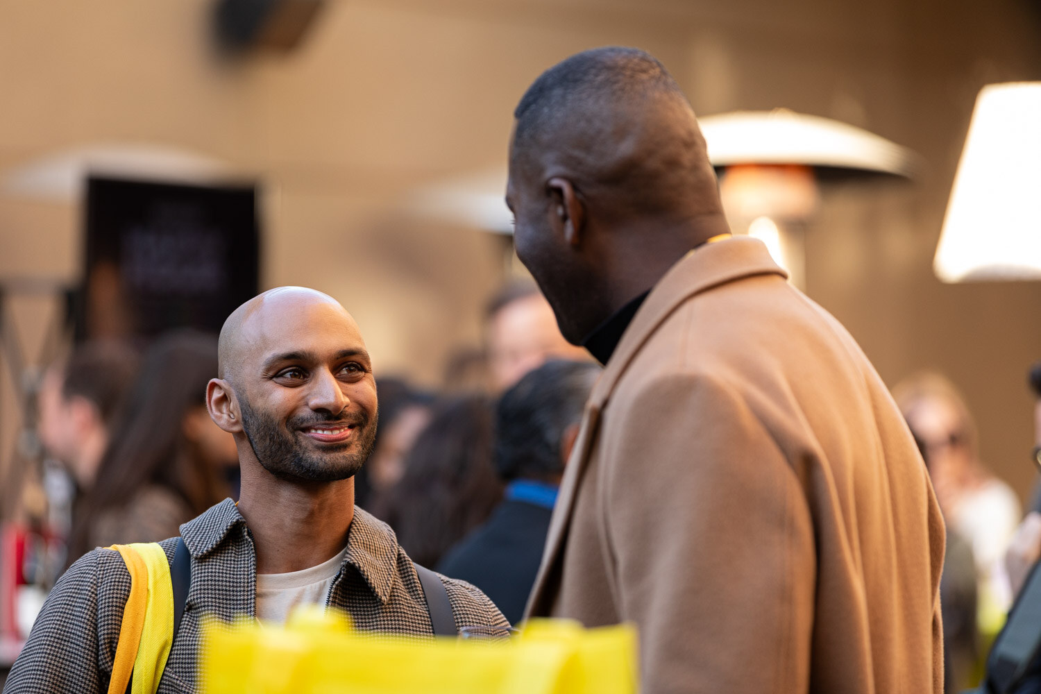 A smiling man talking with another attendee in front of a yellow tote bag.