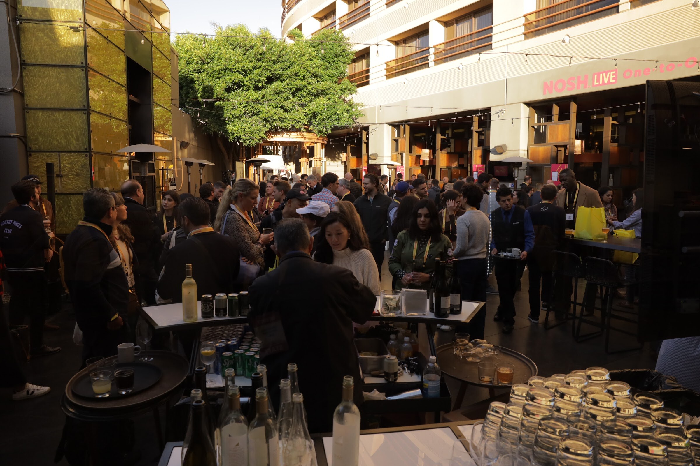 A crowded outdoor courtyard below a pink NOSH LIVE sign with a bar in the foreground.