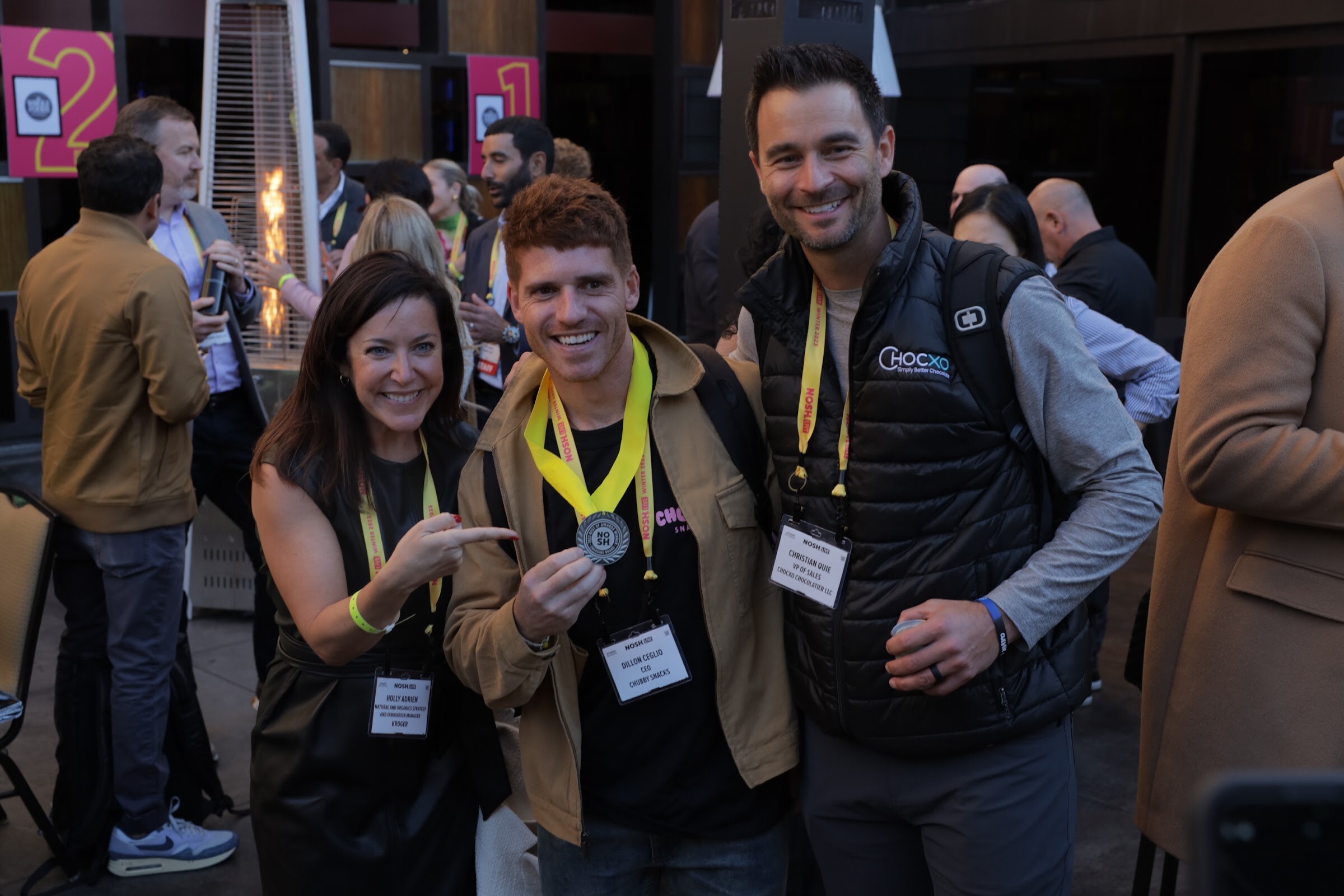 A man and woman posing with a medal on a lanyard in a crowded outdoor reception.