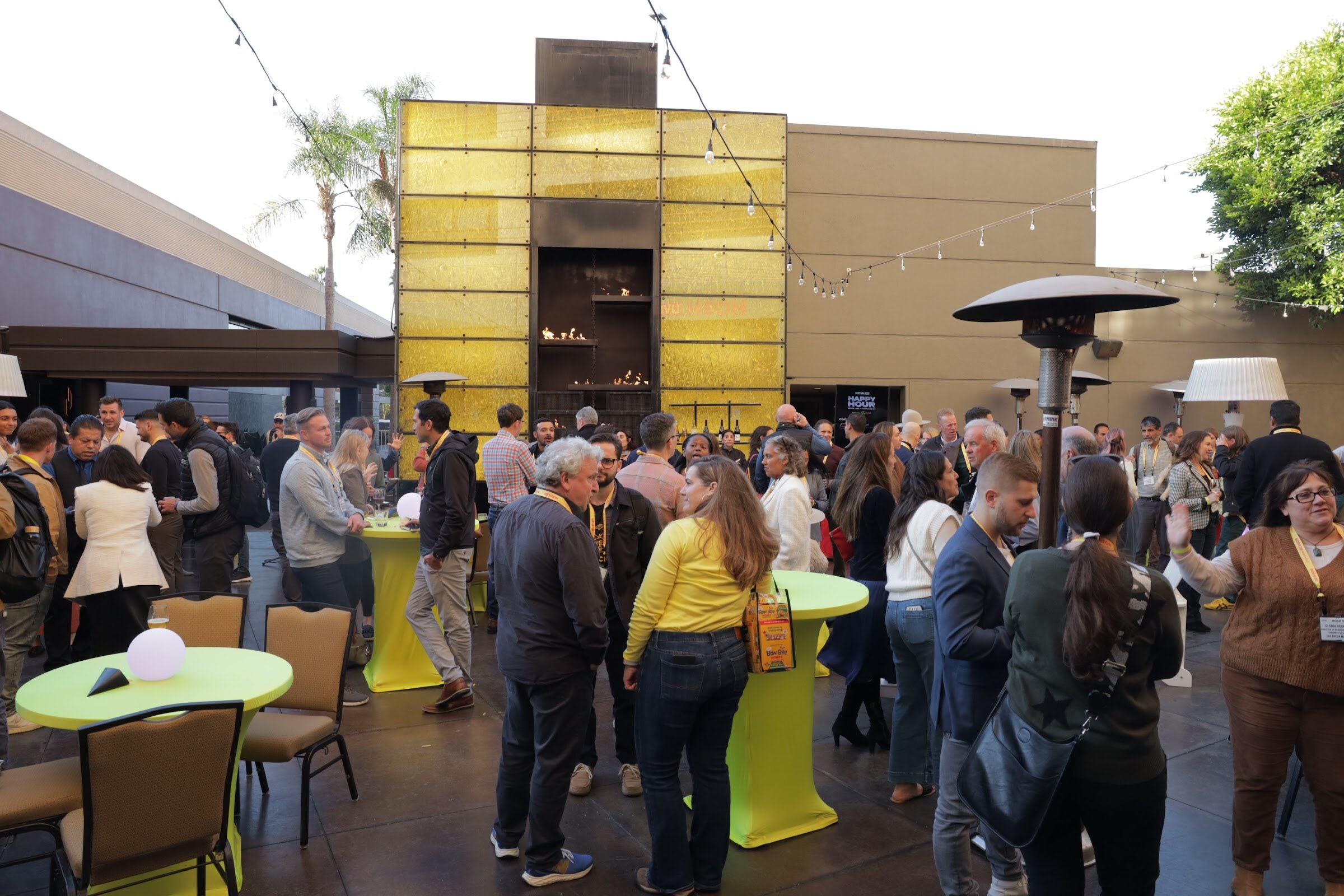 A courtyard with attendees standing around yellow-green high tables in front of a yellow tiled wall.
