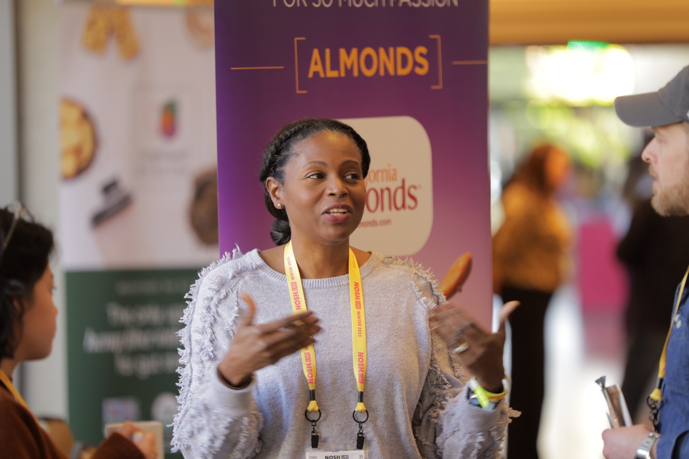 A woman with a lanyard gestures while talking, with a purple Almonds banner behind her.