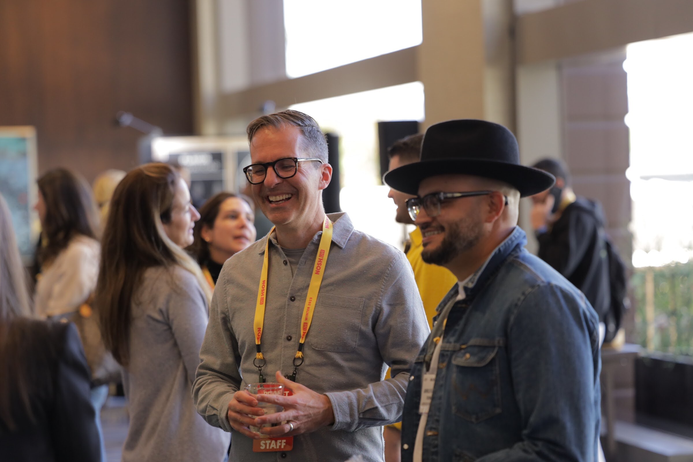 Two men with lanyards smiling in a crowded lobby near large windows.