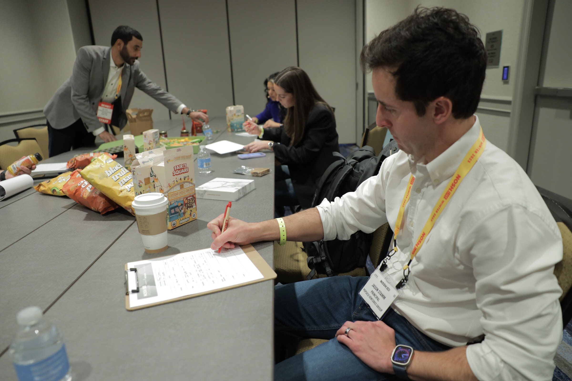 Several judges seated at a table with clipboards, snack packages and a green sample box.