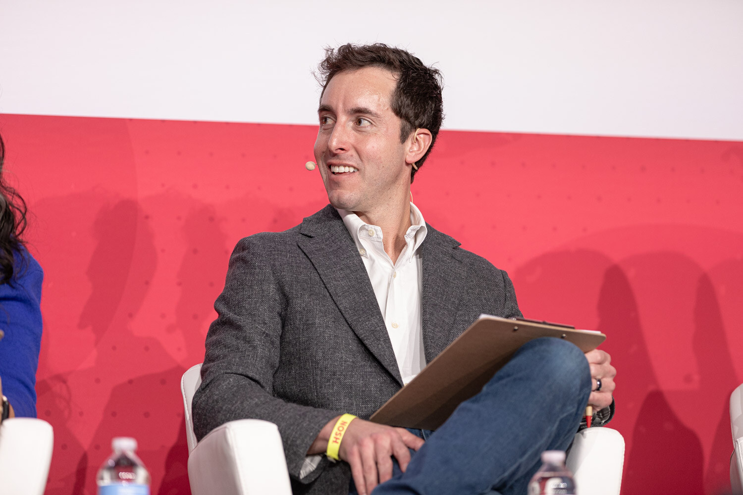 A man in a gray blazer holding a clipboard, seated on stage and looking toward the presenter.
