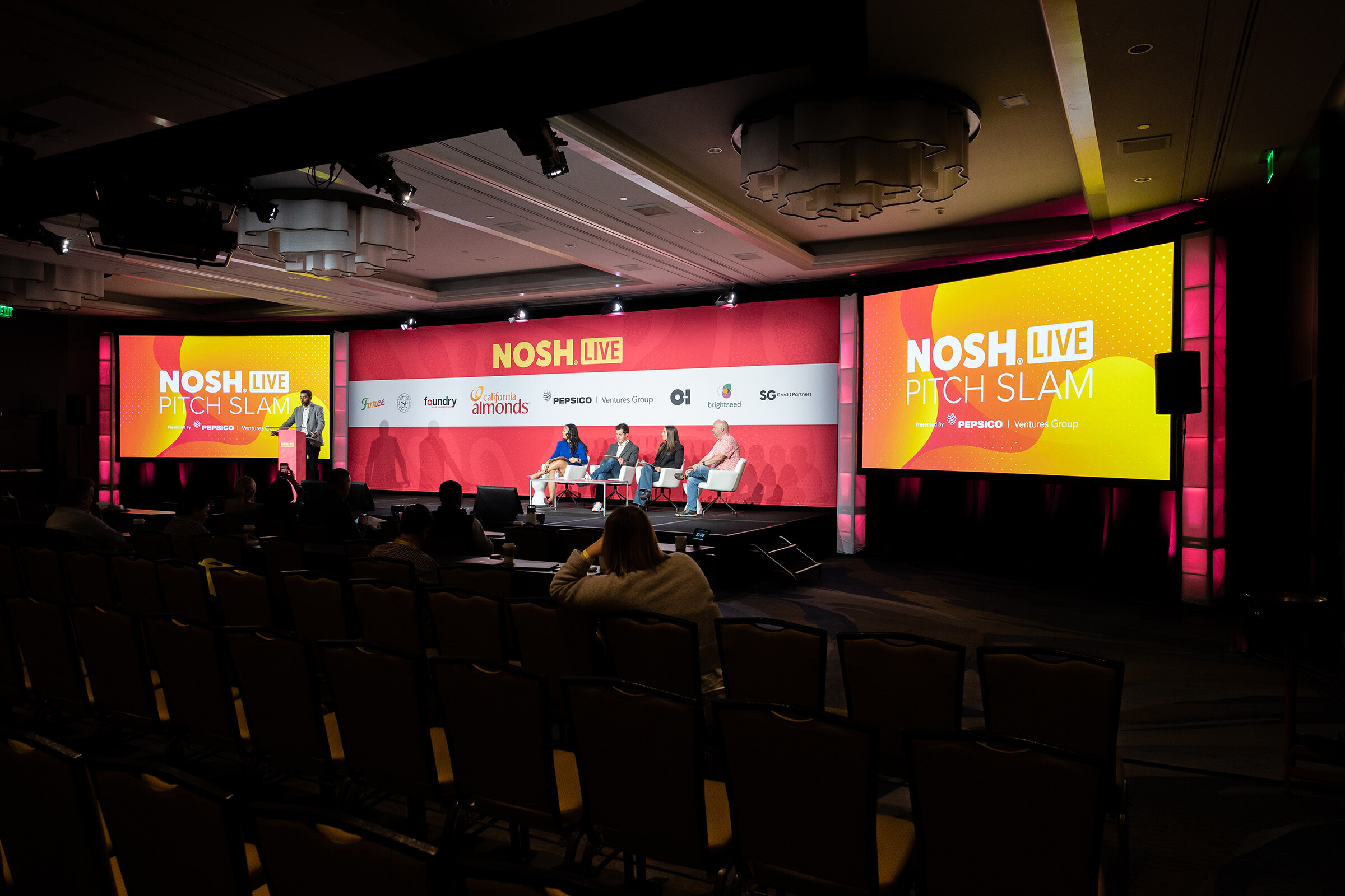 A dark ballroom stage with NOSH LIVE Pitch Slam screens and a row of judges seated on white chairs.