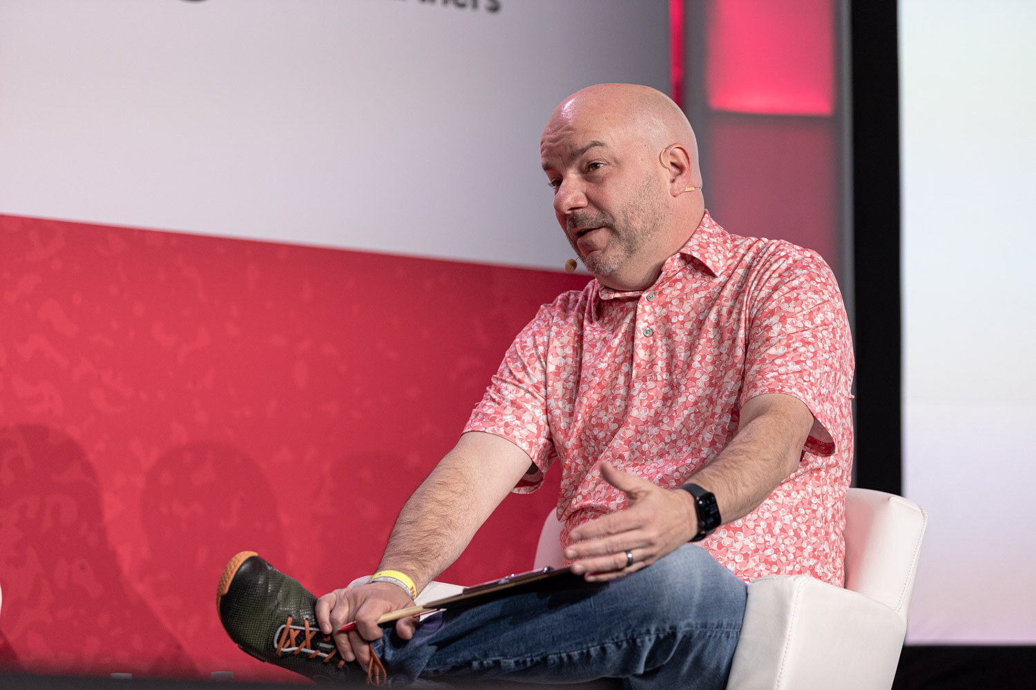 A man in a pink floral shirt seated on stage with a clipboard and one hand extended.