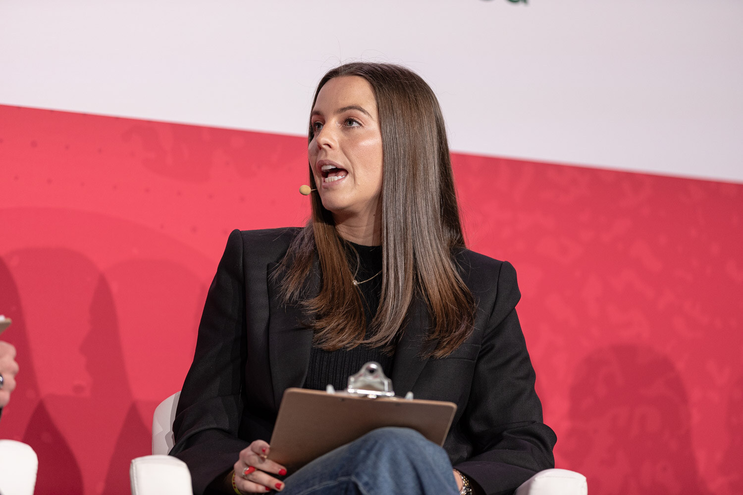 A woman in a black blazer holding a clipboard speaking on stage.