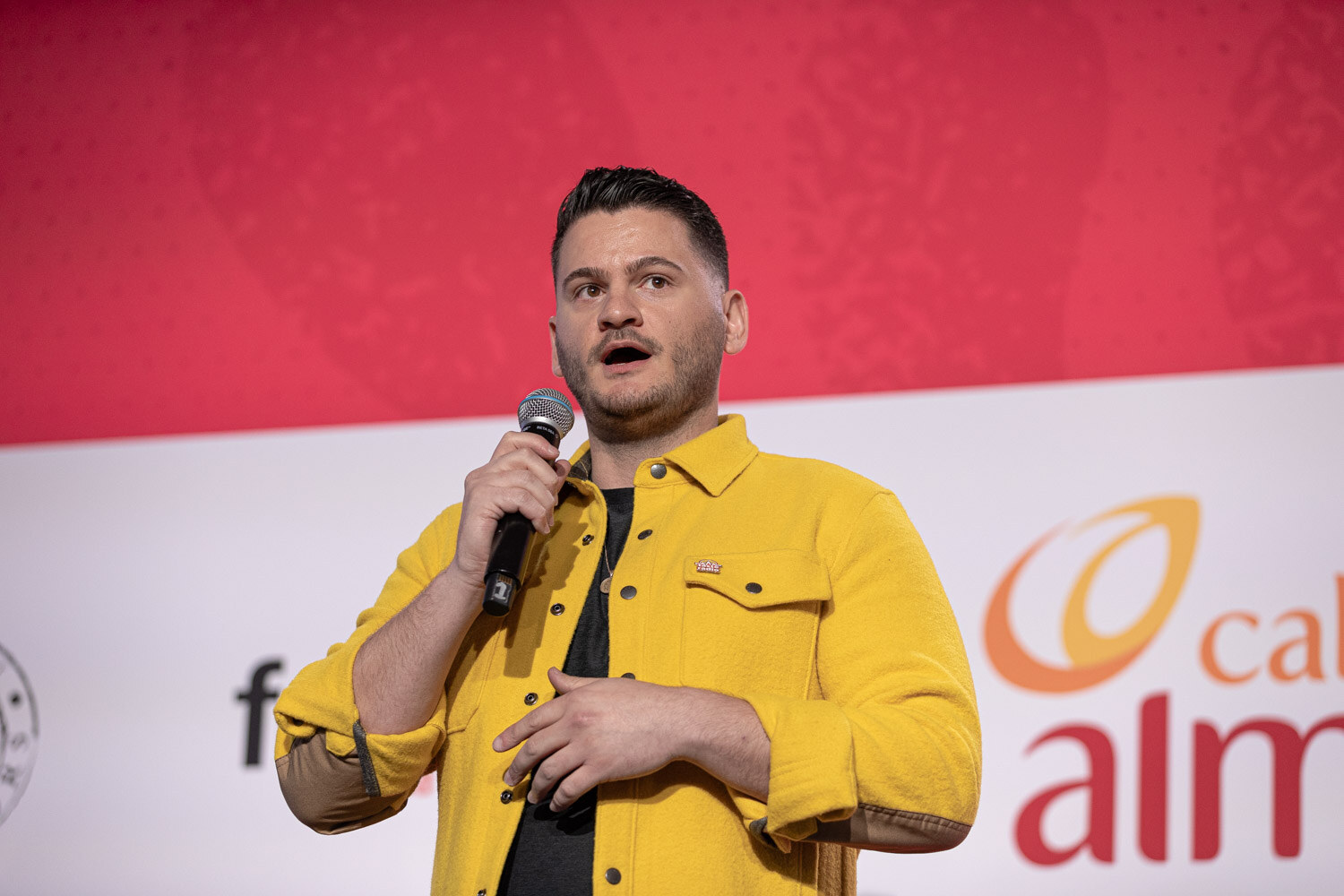A man in a yellow jacket speaking into a microphone in front of the NOSH LIVE stage backdrop.