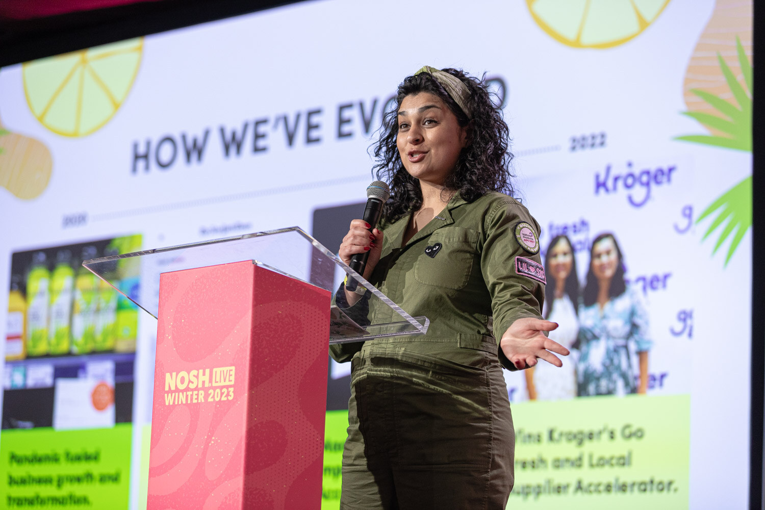 A woman in an olive jumpsuit at a podium with a slide titled How We've Evolved and a Kroger logo.