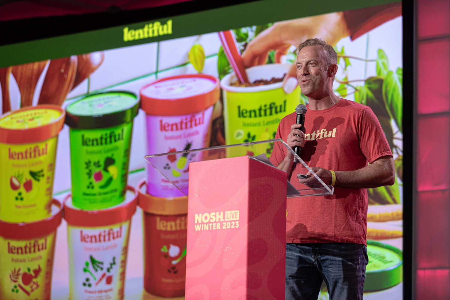 A man in a red Lentiful t-shirt speaking into a microphone in front of a slide of lentil cups.