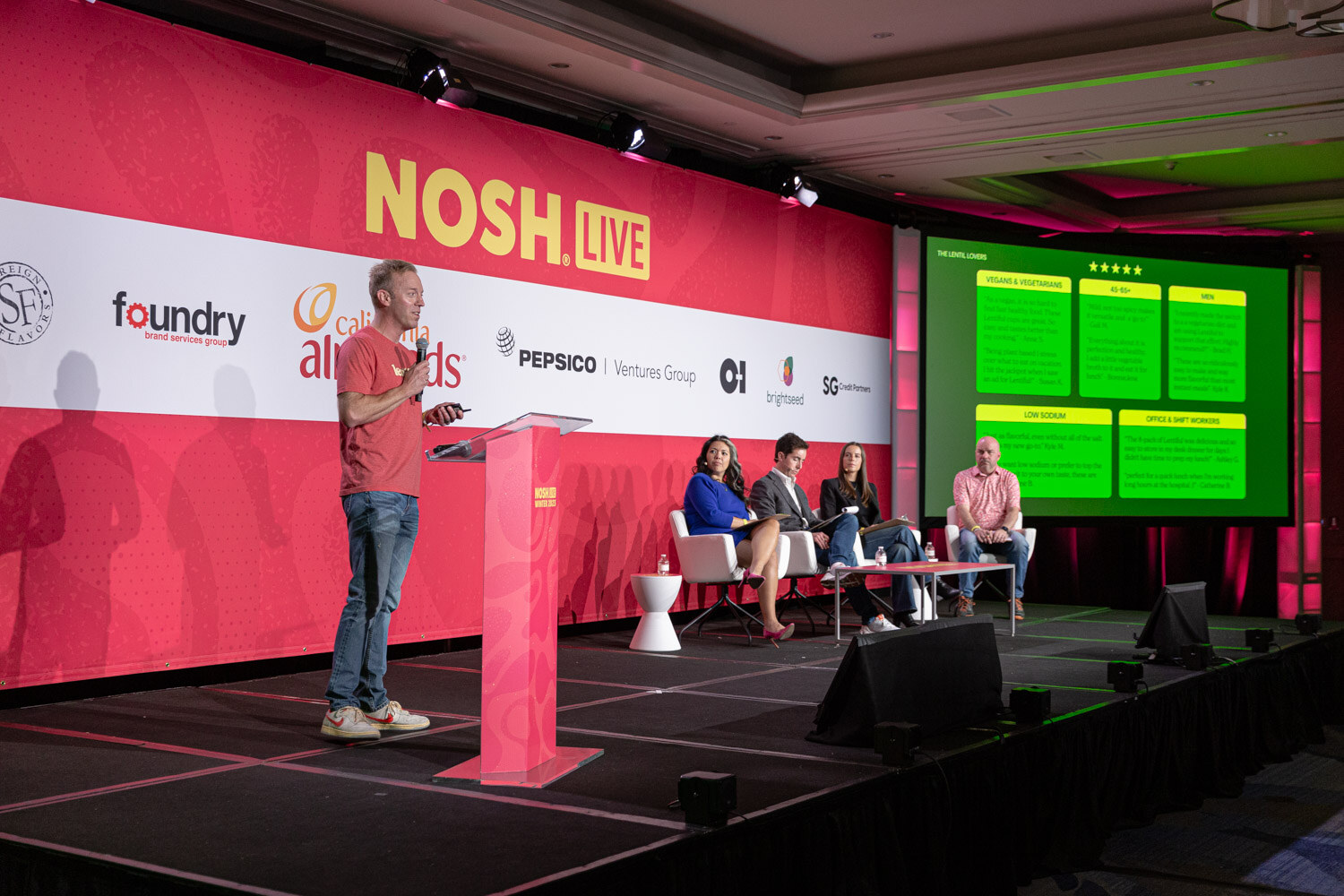 A man in a red shirt at a podium with four judges seated on stage and a green slide on the screen.
