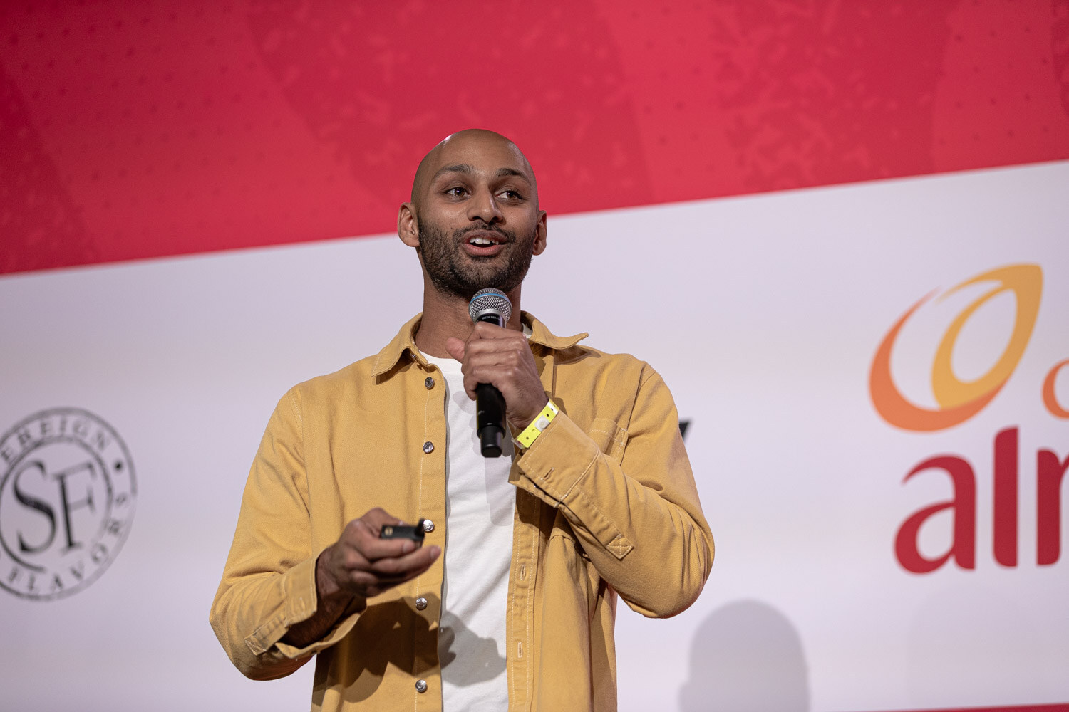 A man in a mustard shirt holding a microphone and clicker in front of the stage backdrop.